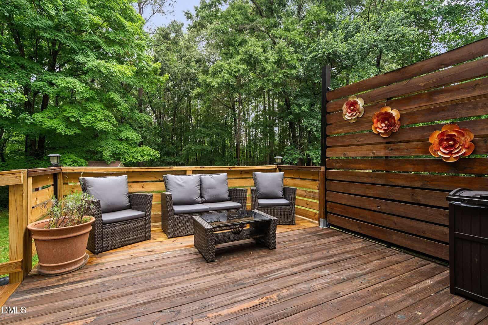 A wooden deck with outdoor wicker seating, gray cushions, a glass coffee table, a large potted plant, and a privacy screen decorated with three flower sculptures, surrounded by green trees.