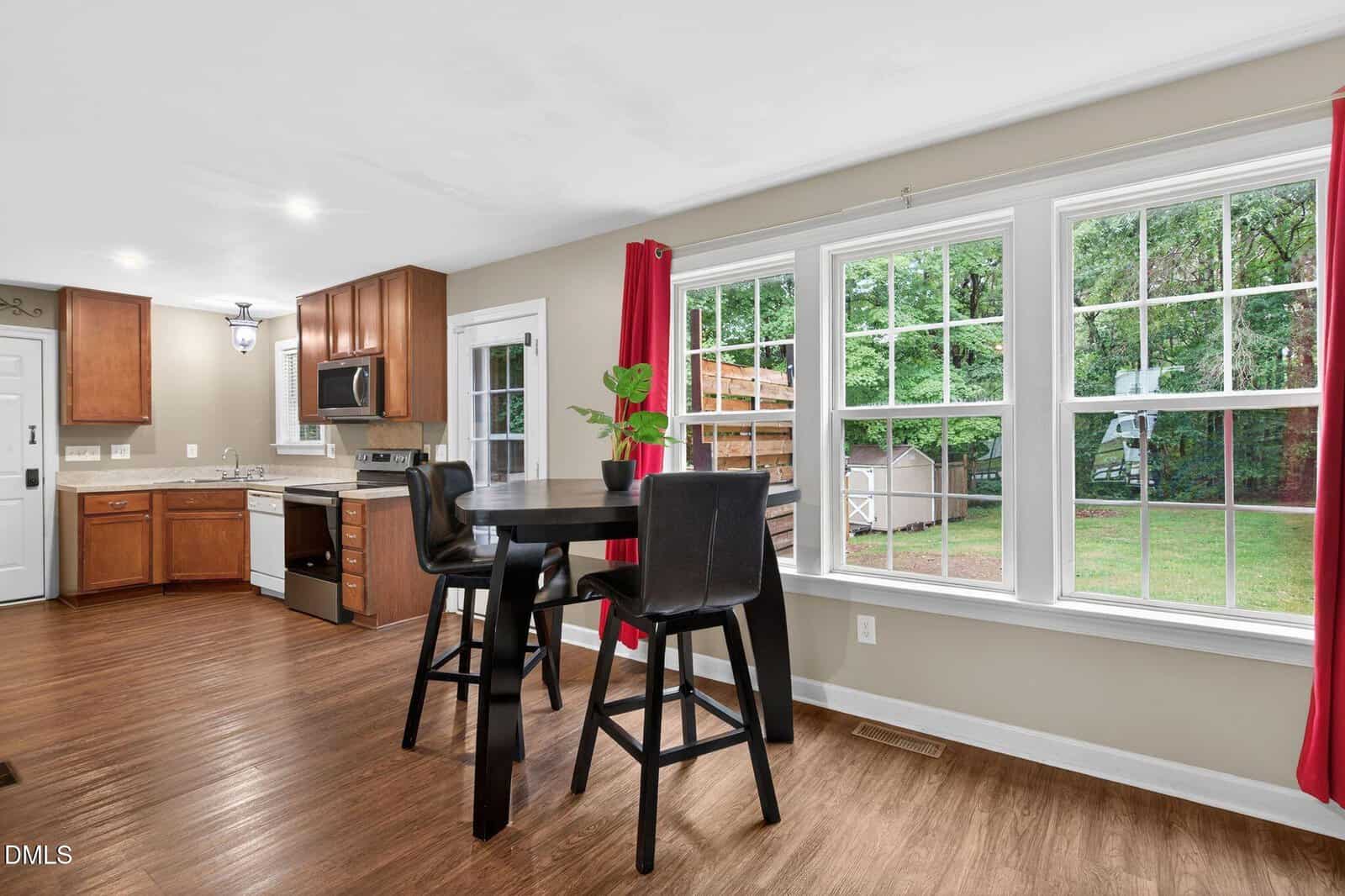 Bright kitchen with wood cabinets, stainless steel appliances, a round dining table with four black chairs near large windows, and views of a green backyard with a playset and shed.