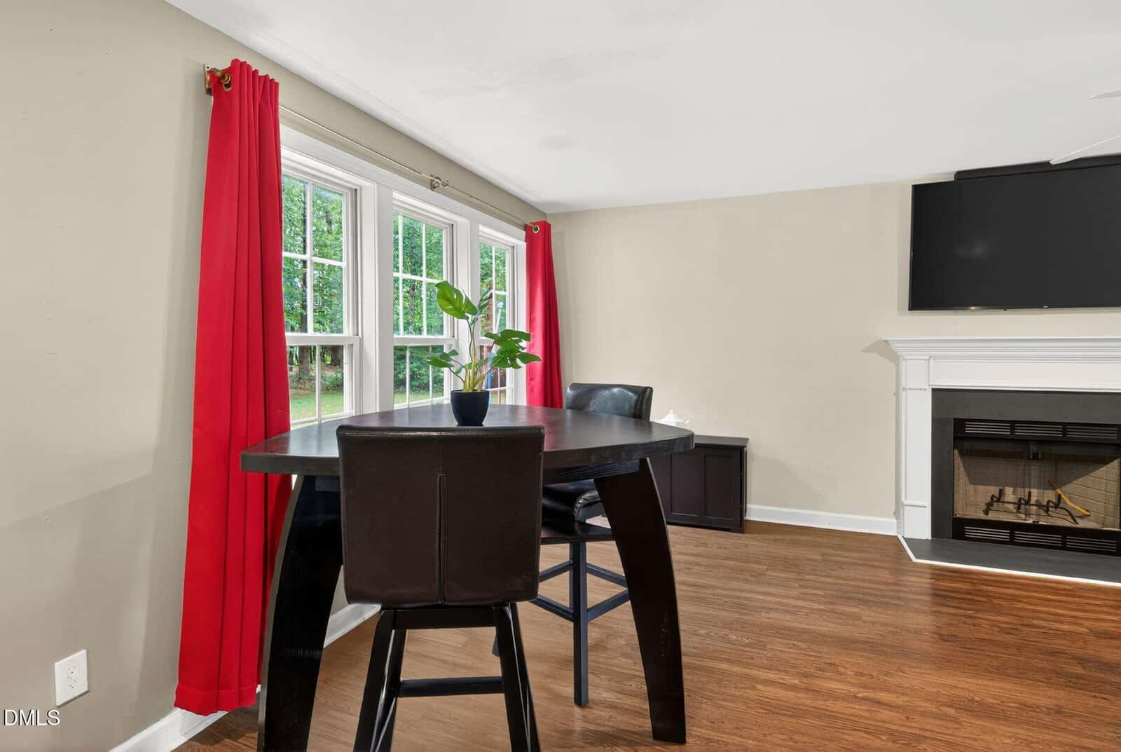 A dining area with a dark wood table, two chairs, a plant centerpiece, red curtains, large windows, hardwood floor, a fireplace, and a wall-mounted TV.