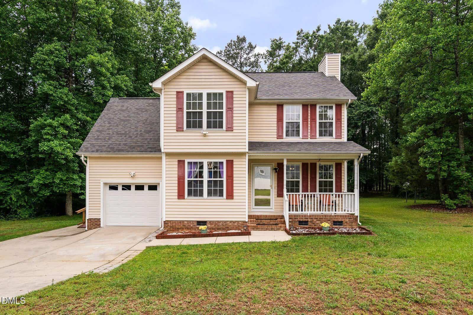 Charming two-story beige house at 115 Cornerstone Drive, Franklinton, with red shutters, a front porch, and single-car garage. Surrounded by green grass, landscaped flowerbeds, and large trees. A driveway leads up to the garage.