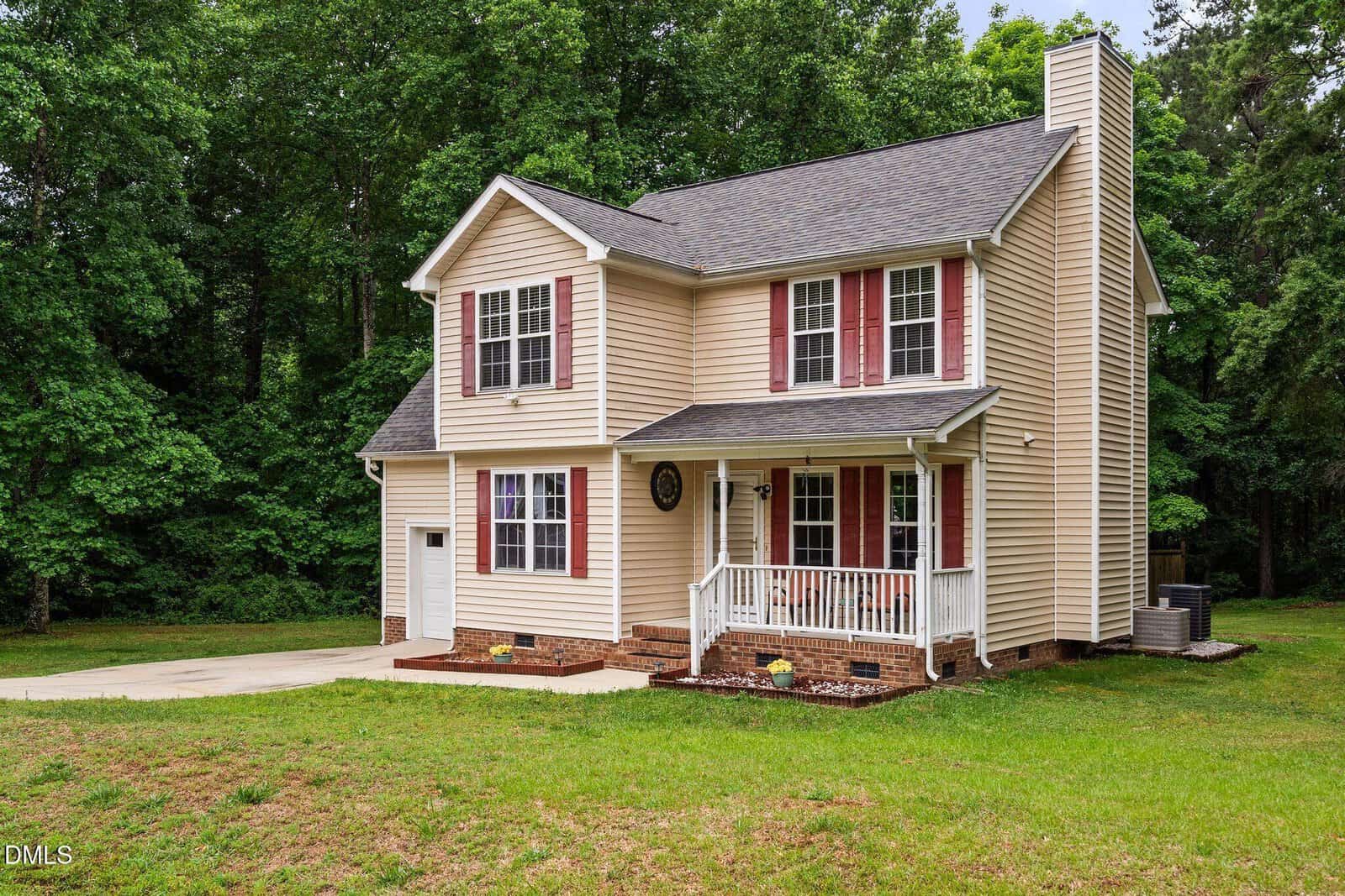 Two-story beige house with white trim, red shutters, a front porch, and a brick foundation, situated on a grassy yard with a driveway and surrounded by green trees.