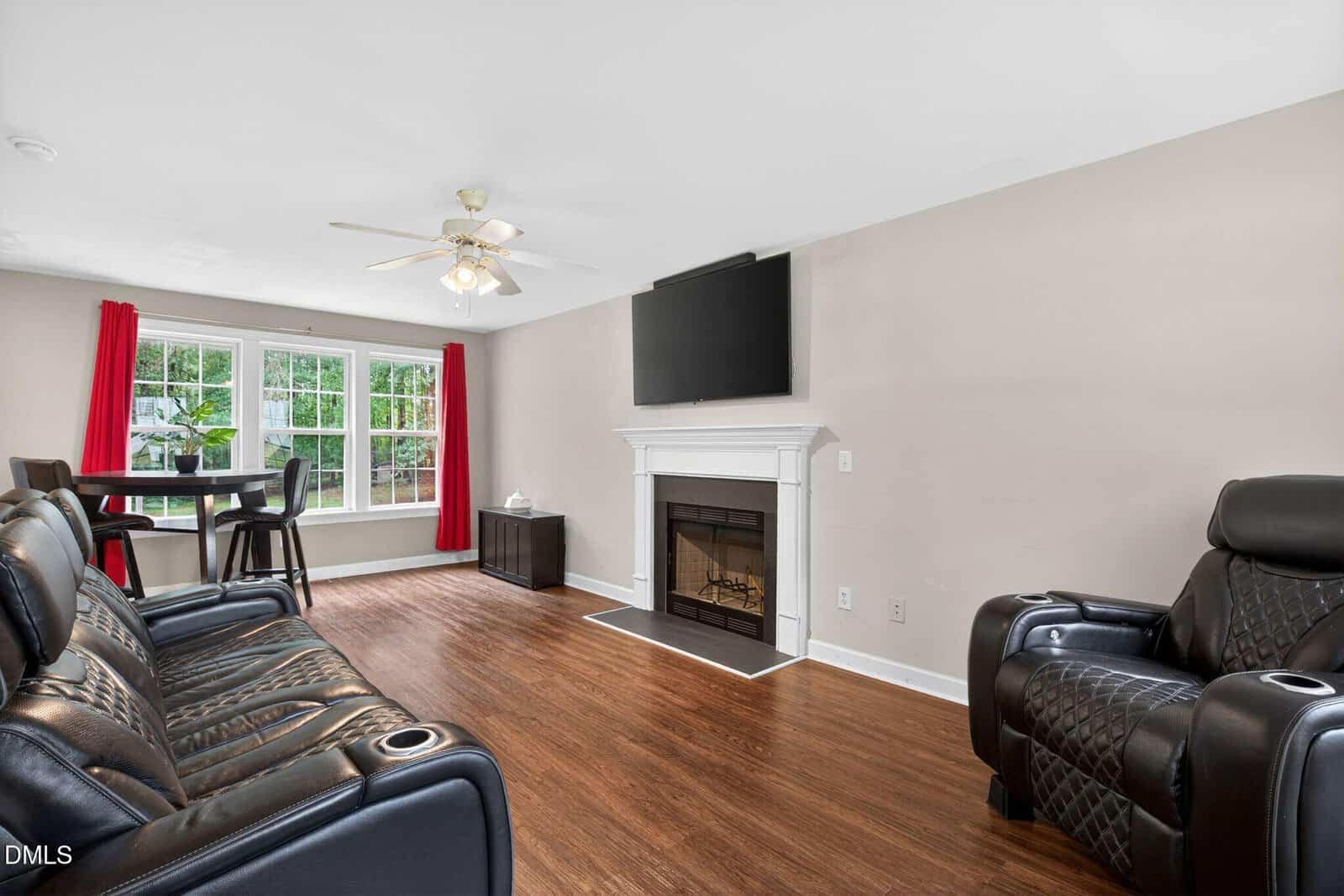 Living room with black leather furniture, a white fireplace with a TV above it, wood flooring, a dining table by large windows with red curtains, and neutral-colored walls.