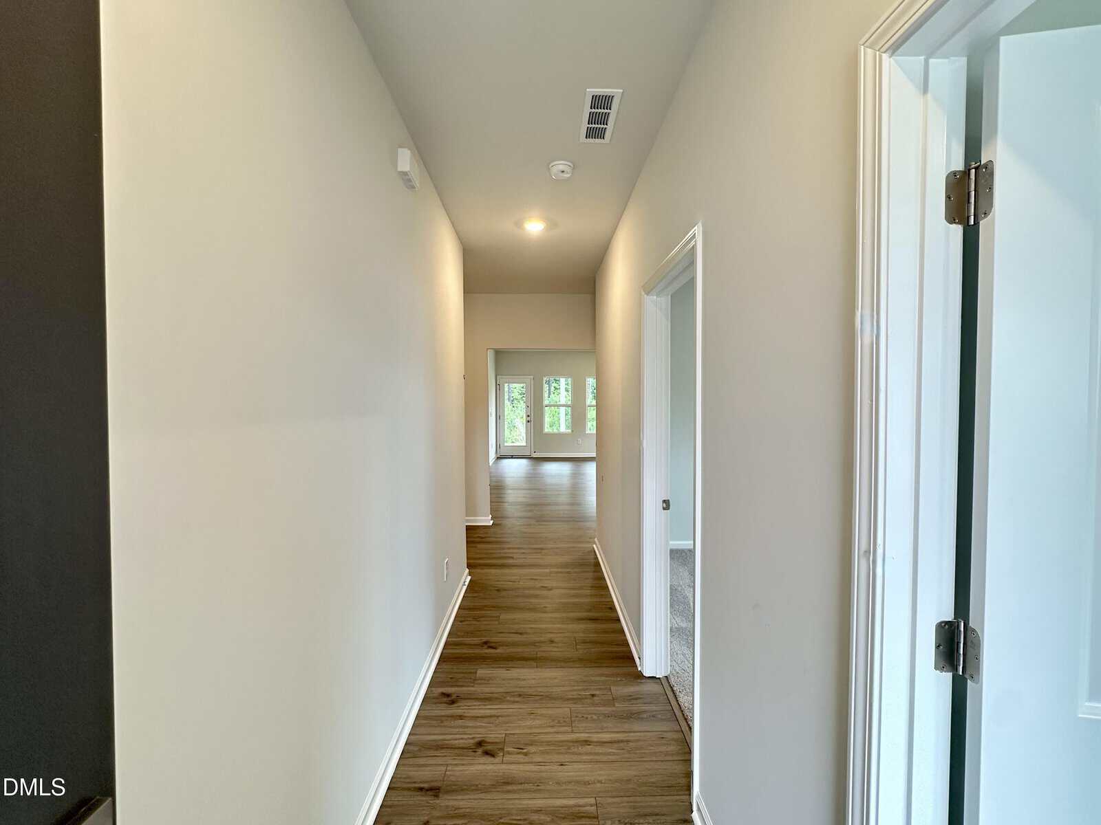 A hallway with light-colored walls and wood flooring, leading to a room with large windows. There are open doorways on the right side and a ceiling vent visible.