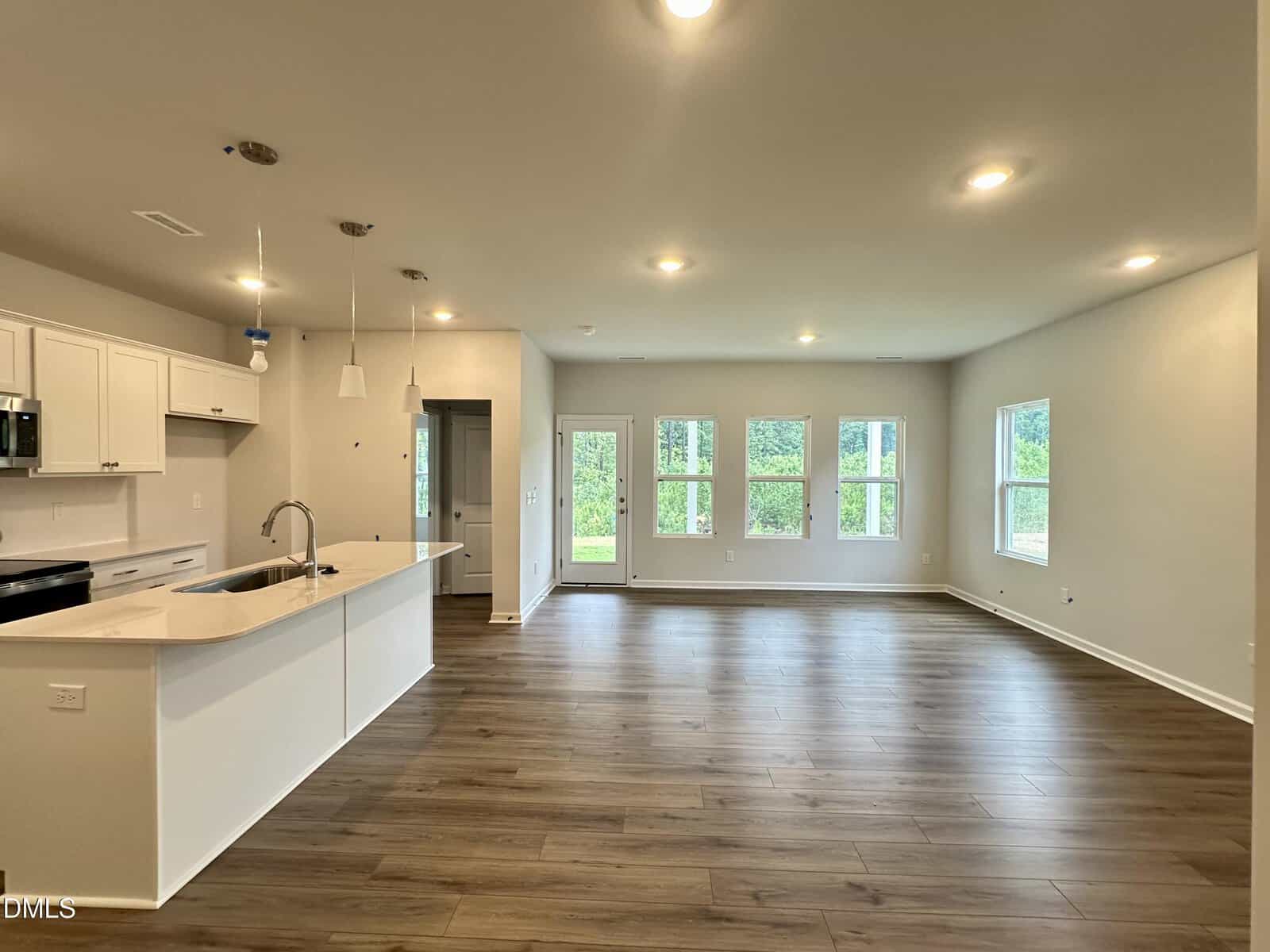 Open-concept kitchen and living area with wood floors, white cabinets, island sink, and large windows letting in natural light. Walls are light-colored, and the room appears spacious and modern.