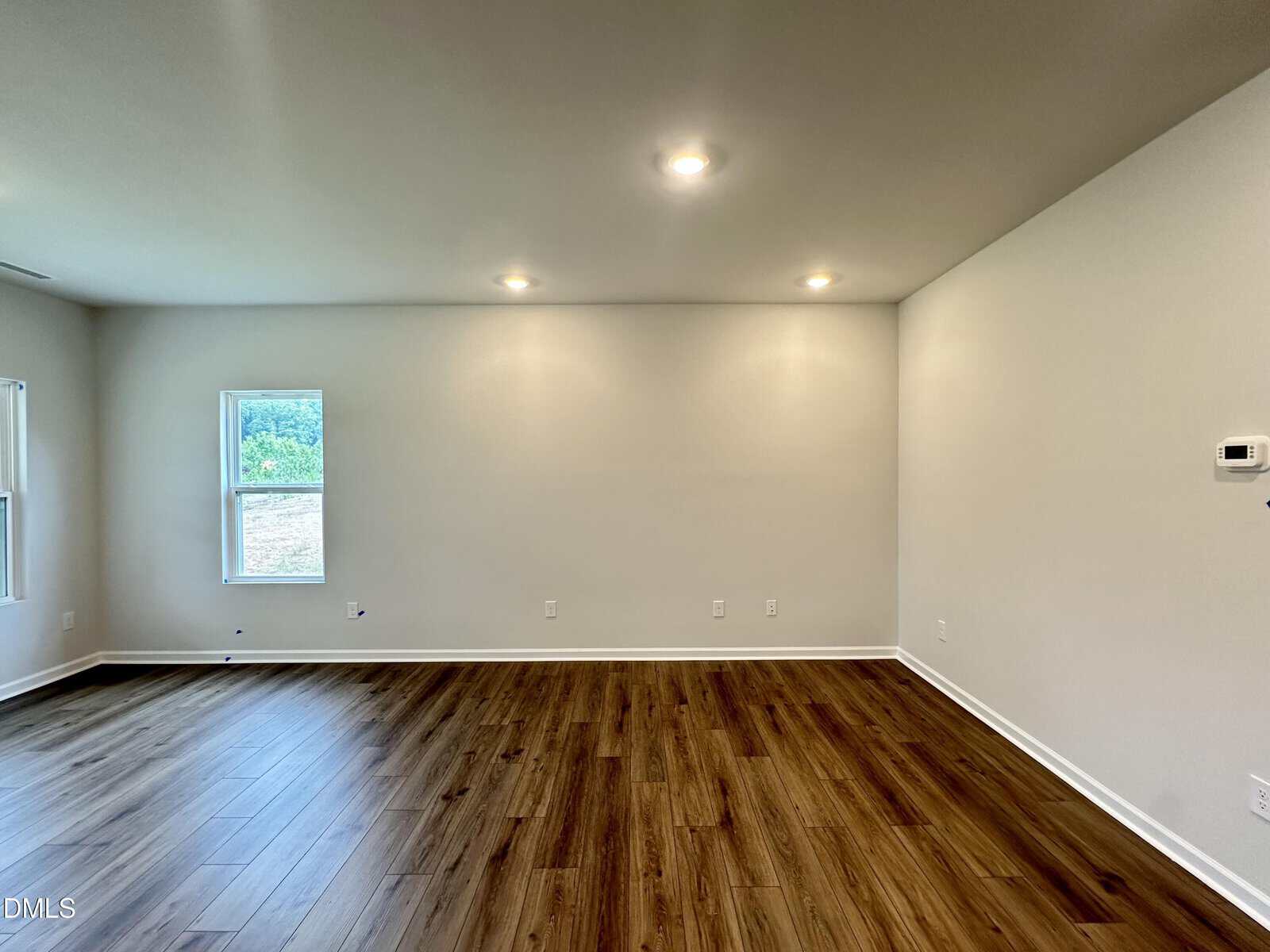 Empty room with light-colored walls, wood flooring, three ceiling lights, two windows on the left wall, and a thermostat on the right wall. The room has a clean and modern appearance.
