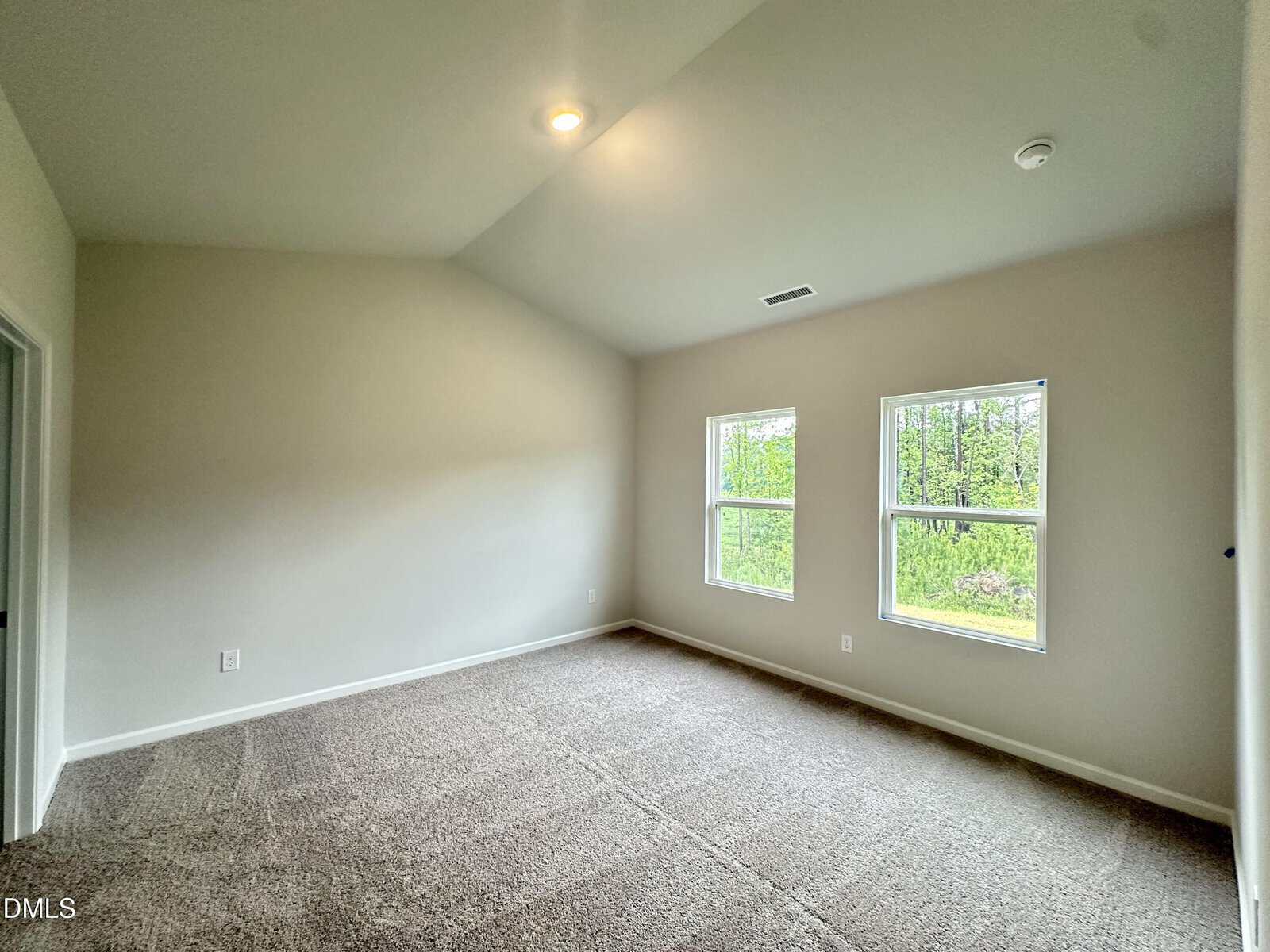 Empty carpeted room with vaulted ceiling, two windows showing green trees outside, neutral-colored walls, and a single ceiling light. Doorway visible on the left side.