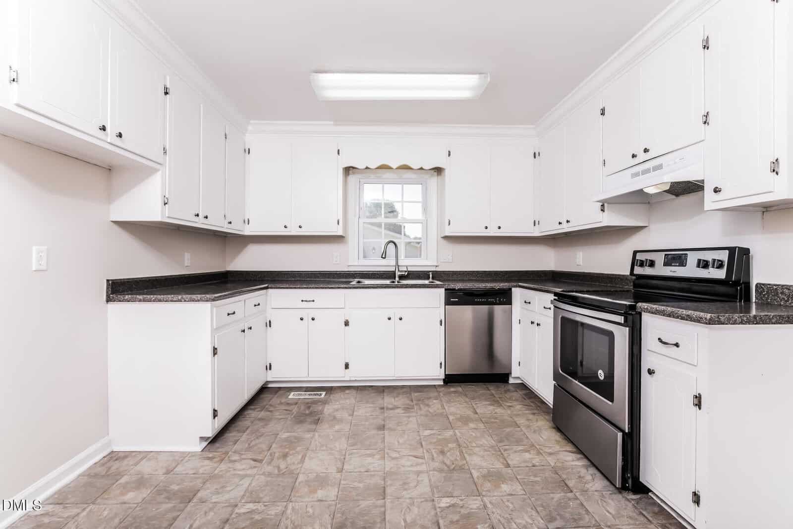 Bright kitchen with white cabinets, dark countertops, stainless steel appliances, electric stove, dishwasher, double sink under a window, and gray tile flooring. Walls are light, and lighting is fluorescent.