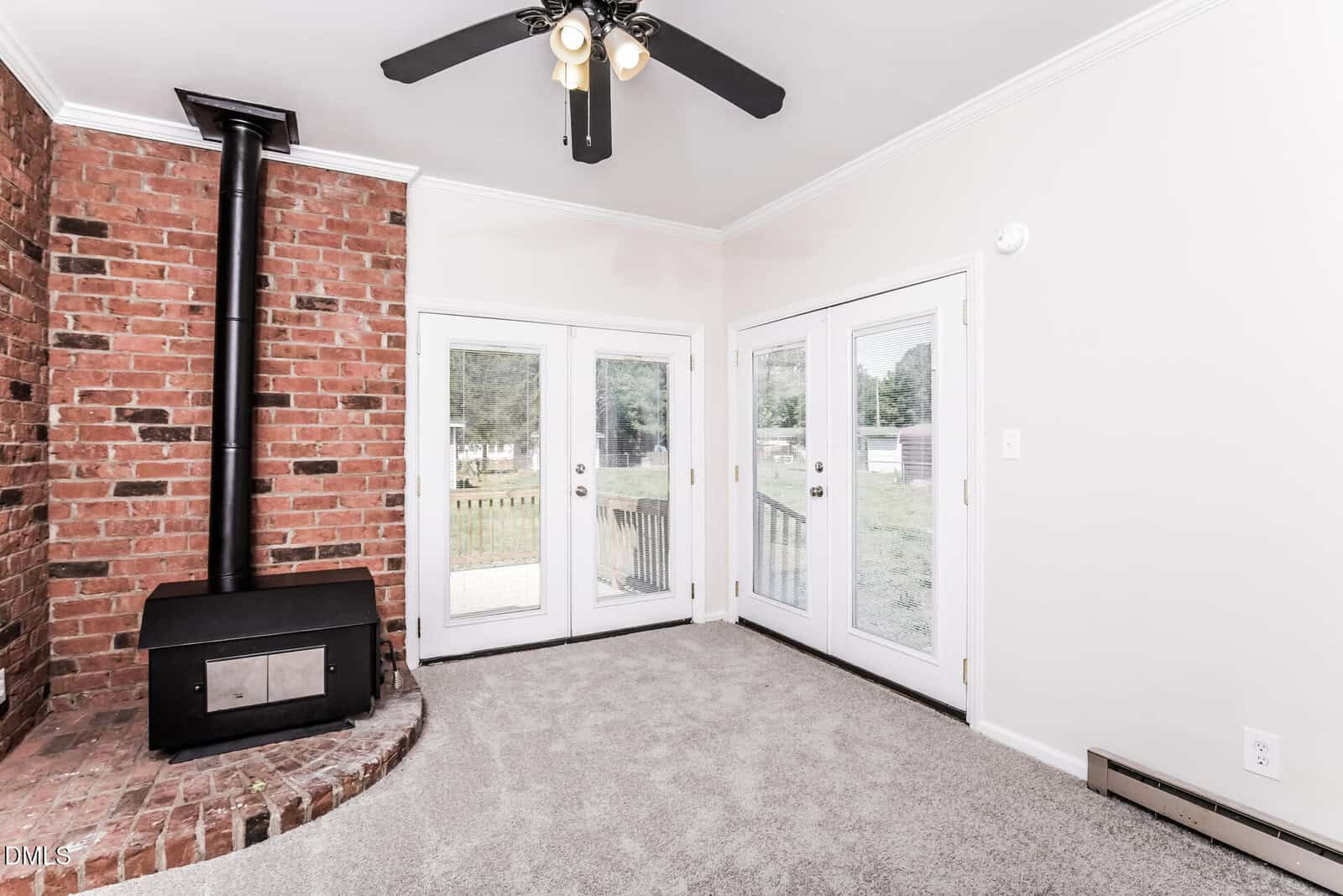 A bright room with light walls and carpet, featuring a black wood stove set against a red brick corner, a ceiling fan, and double French doors opening to a backyard with green grass.