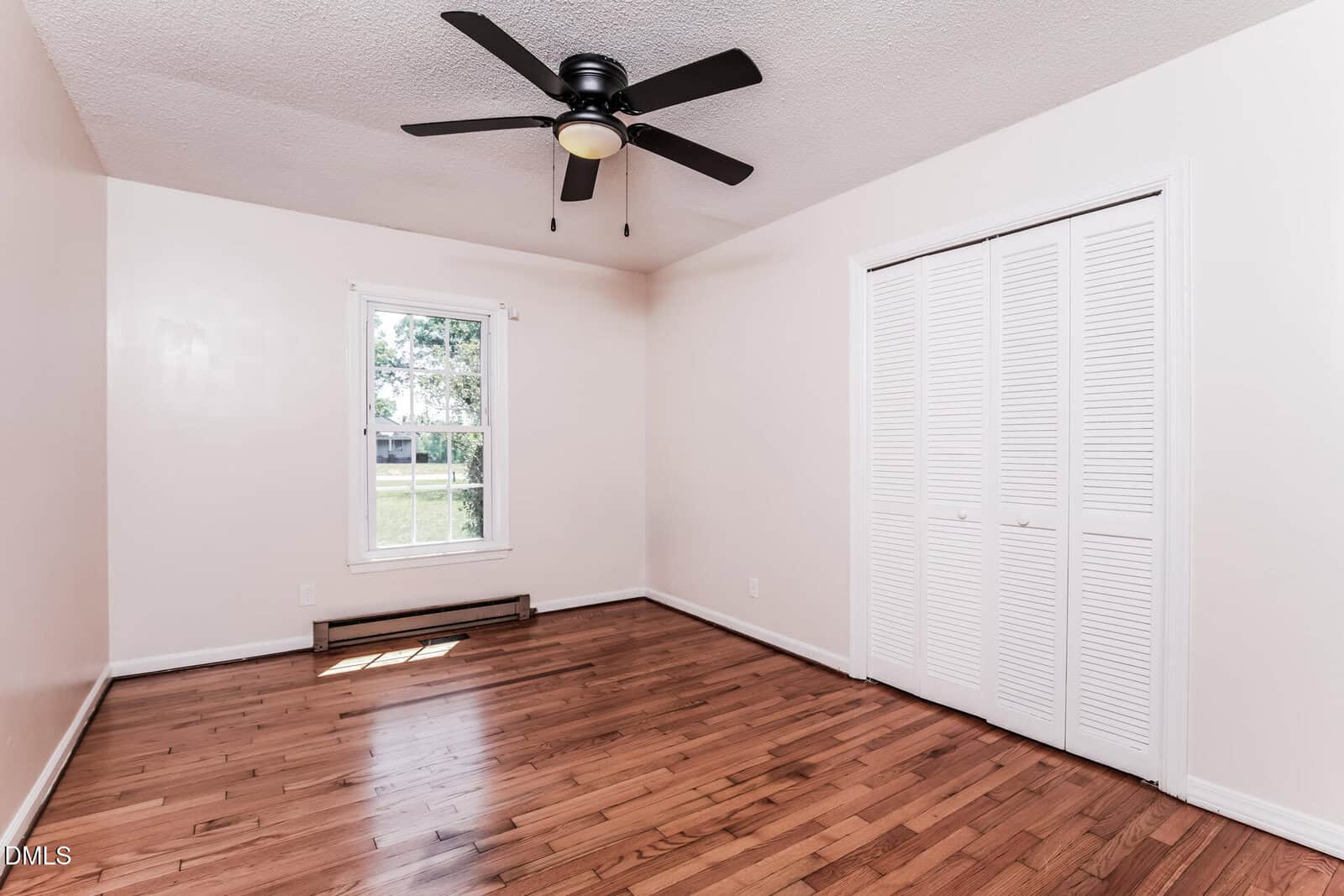 A bright, empty room with light-colored walls, hardwood floors, a ceiling fan with four blades, a window letting in sunlight, and a white double-door closet.