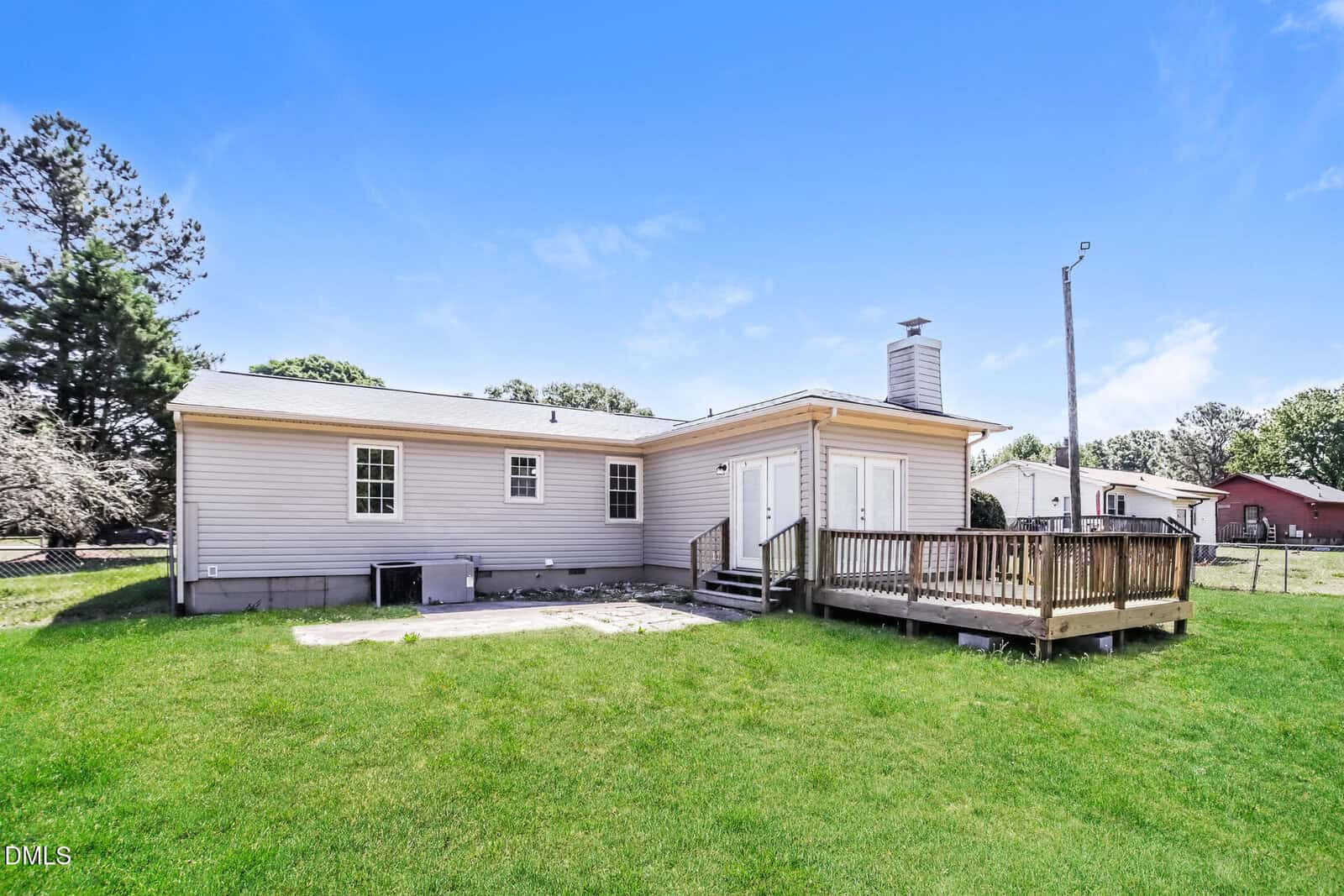 A single-story house with light siding, multiple windows, a small deck with steps, and a grassy fenced backyard under a clear blue sky.