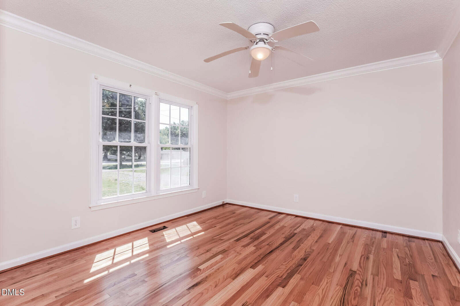 Empty room with light-colored walls, wooden floor, large double window letting in natural light, a white ceiling with crown molding, and a ceiling fan with a light fixture.