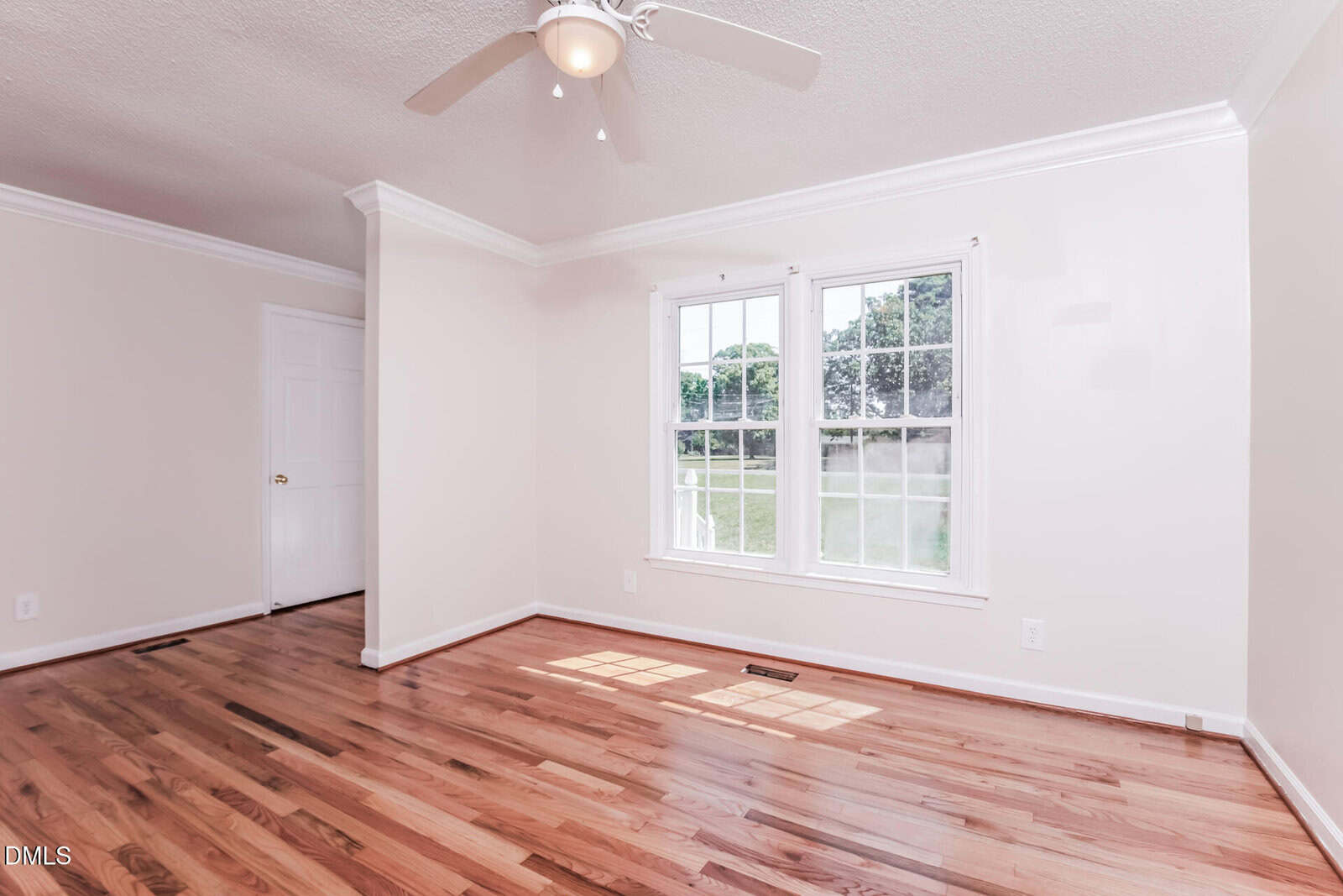 Empty room with light hardwood floors, white walls, a ceiling fan, large window with outdoor view, white door, and crown molding. Natural light brightens the space.