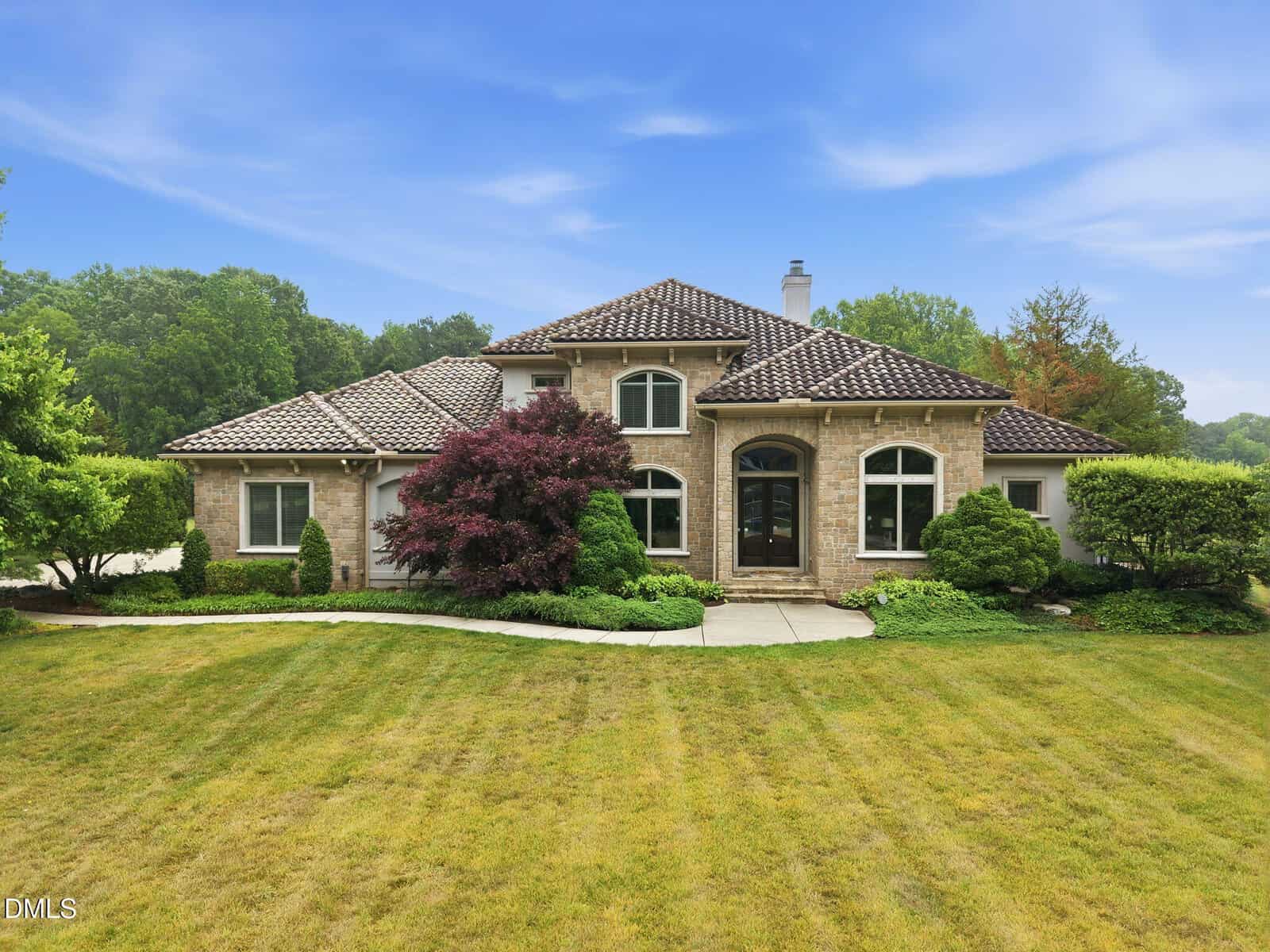 A two-story, tan brick house with a dark tiled roof, arched windows, and manicured landscaping sits on a large, green lawn under a blue sky with scattered clouds.