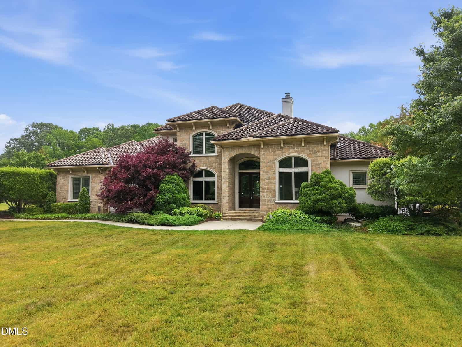 Elegant two-story house with stone exterior, arched windows, and a tiled roof, surrounded by lush green lawn, shrubs, and trees under a clear blue sky.