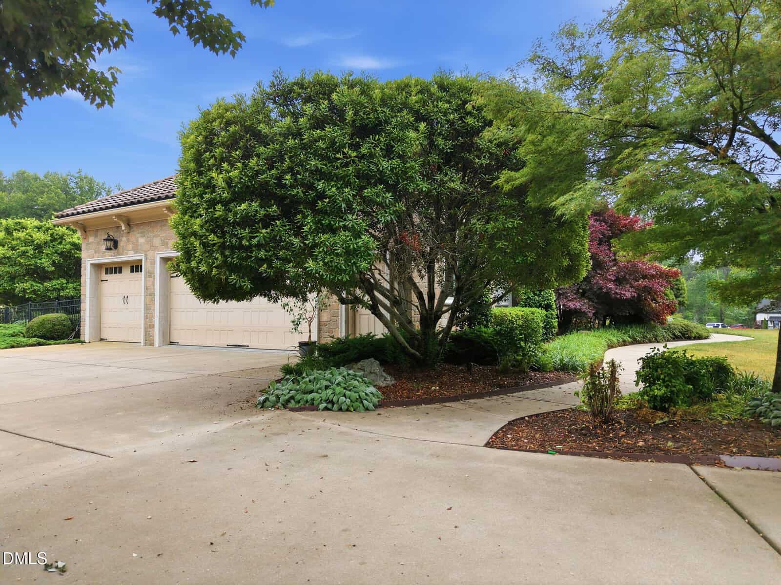 A two-car garage with light-colored doors is partially hidden by a large, leafy tree. The driveway and walkway are bordered by landscaped greenery and flower beds, with a lawn and more trees in the background under a blue sky.
