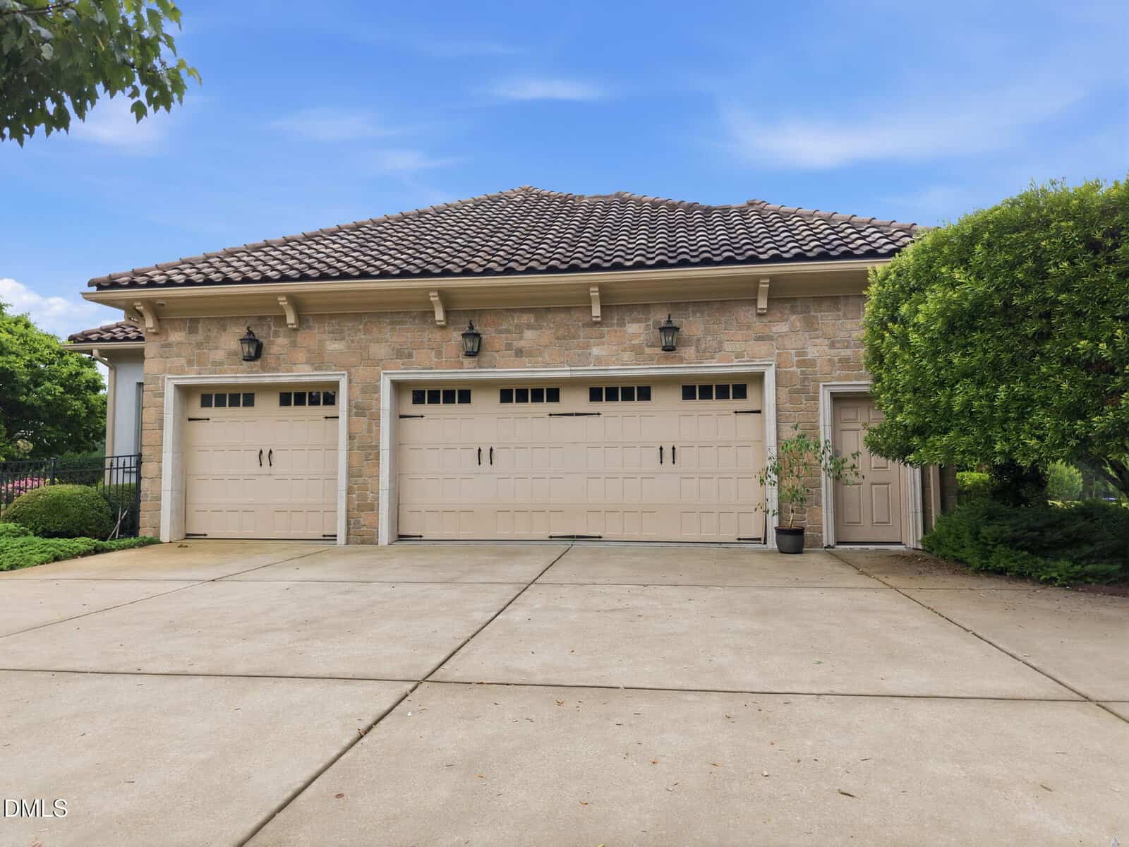 A stone three-car garage with beige doors, lantern-style lights above each door, a tiled roof, and a small potted plant near a side door. The driveway is wide and bordered by lush green trees and shrubs.