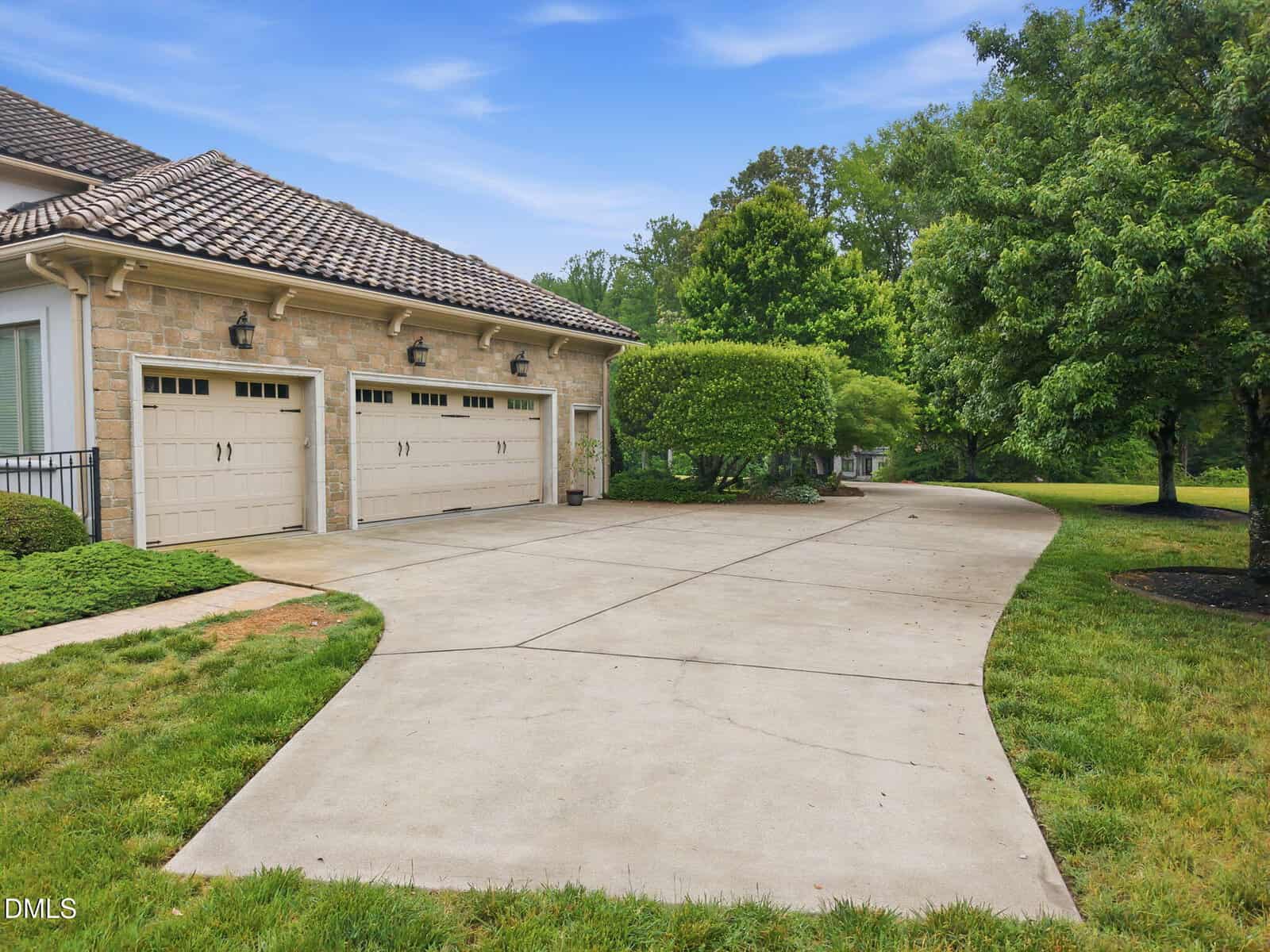 A wide concrete driveway curves toward a three-car garage attached to a house, surrounded by green grass, manicured bushes, and tall trees under a blue sky.