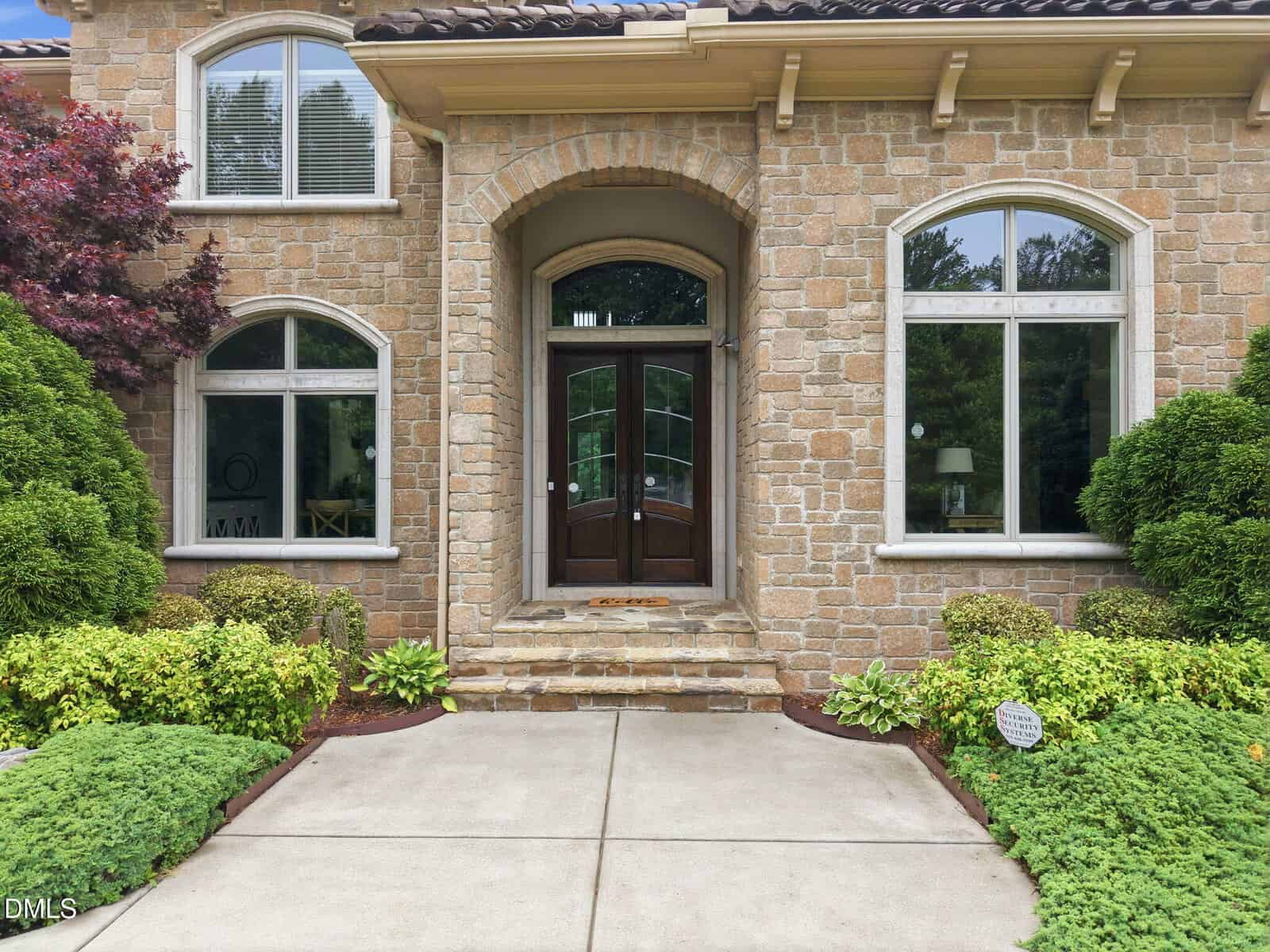 Front entrance of a stone house with large arched wooden door and sidelights, two arched windows, manicured shrubs, and symmetrical landscaping beside a concrete walkway.