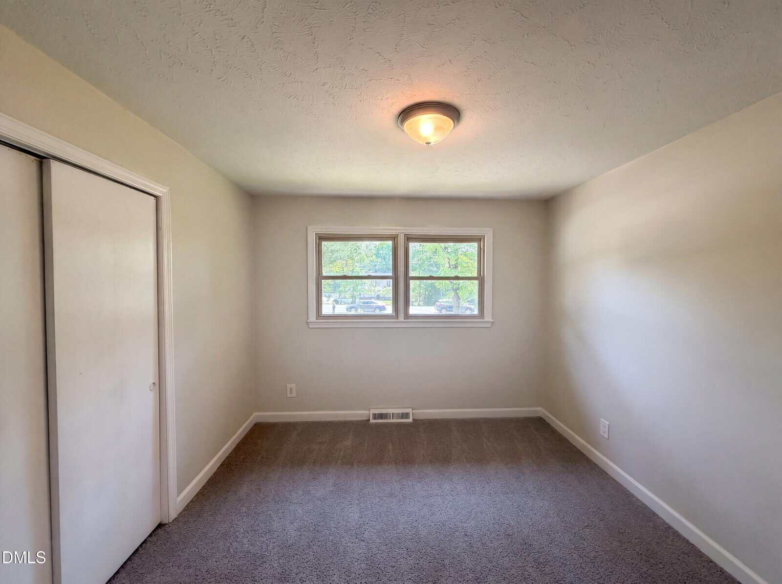 Empty, carpeted room with beige walls, a ceiling light, closed double closet doors on the left, and two windows letting in natural light. There is a floor vent below the windows.