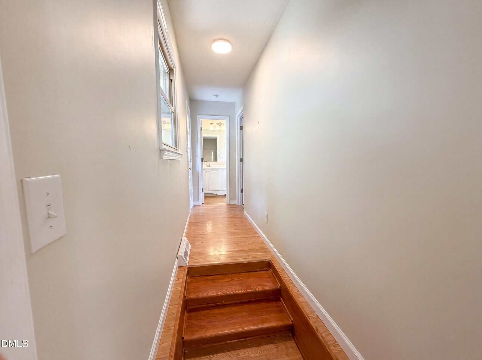 A narrow hallway with wooden floors and a small set of steps leading to a bright room with a white vanity at the end. Pale walls, a window on the left, and a ceiling light brighten the space.