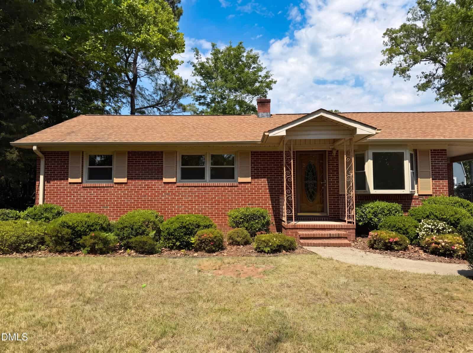 A single-story brick house at 1710 W B Street in Butner features tan shutters and a covered front porch, surrounded by trimmed bushes. The lawn is dry, with trees in the background under a partly cloudy sky.