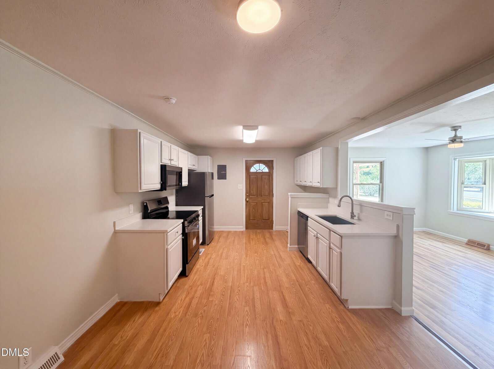 Bright kitchen with light wood flooring, white cabinets, black appliances, a wooden door at the back, and an adjacent open living area with large windows letting in natural light.