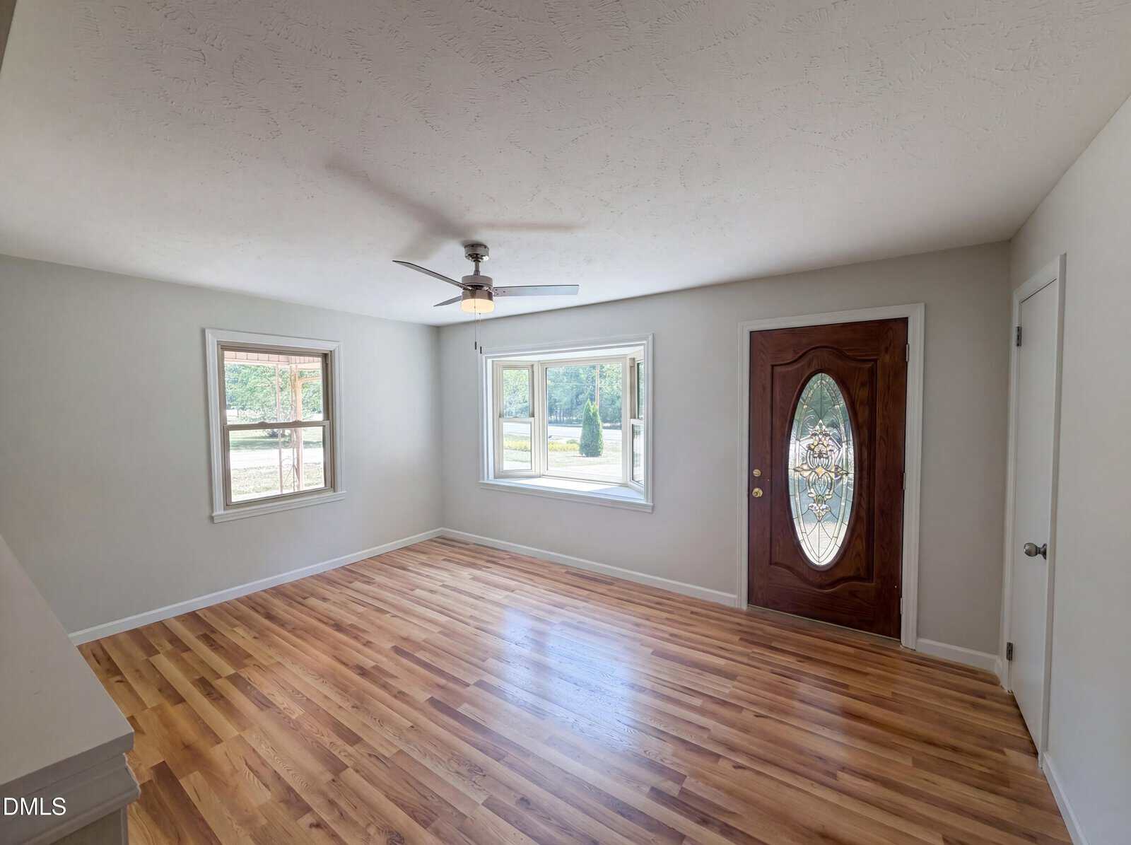 A bright, empty room with light wood flooring, white walls, a ceiling fan, two large windows, and a wooden front door with an oval glass panel. Natural light fills the space.