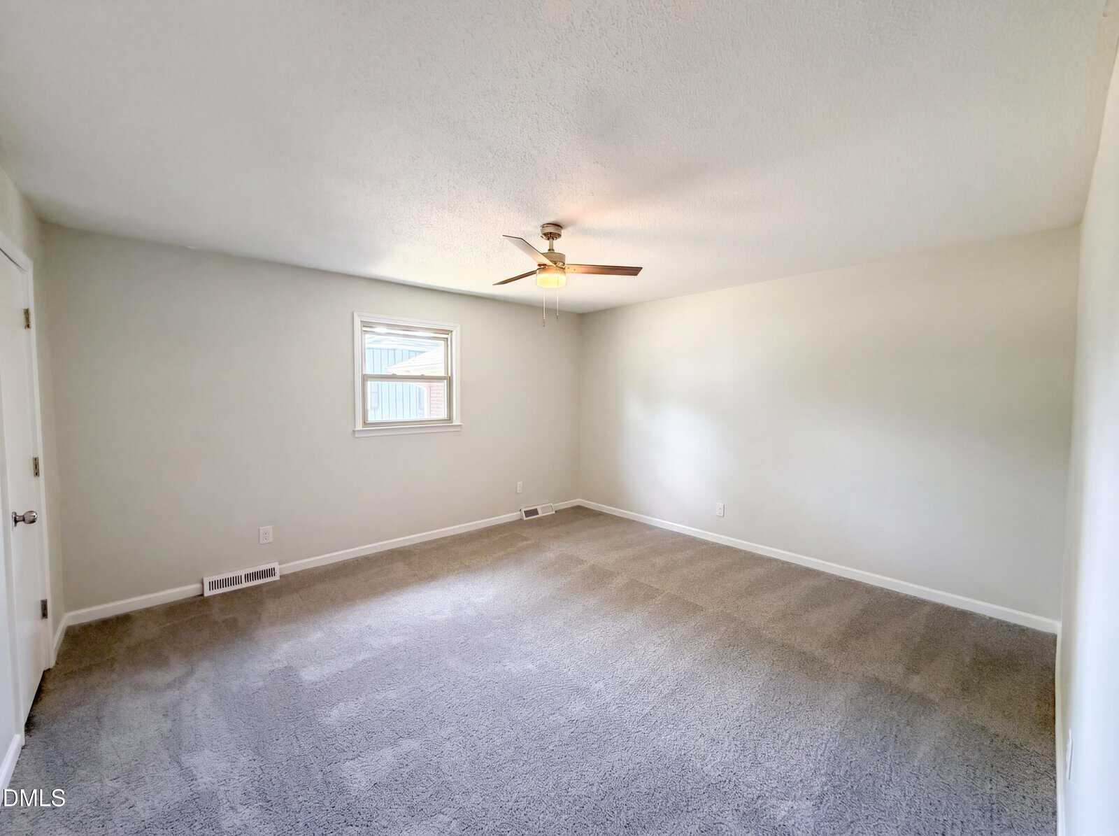 A spacious, empty room with light-colored walls, a gray carpeted floor, a window, and a ceiling fan with a light fixture. There are two closed doors on the left wall.