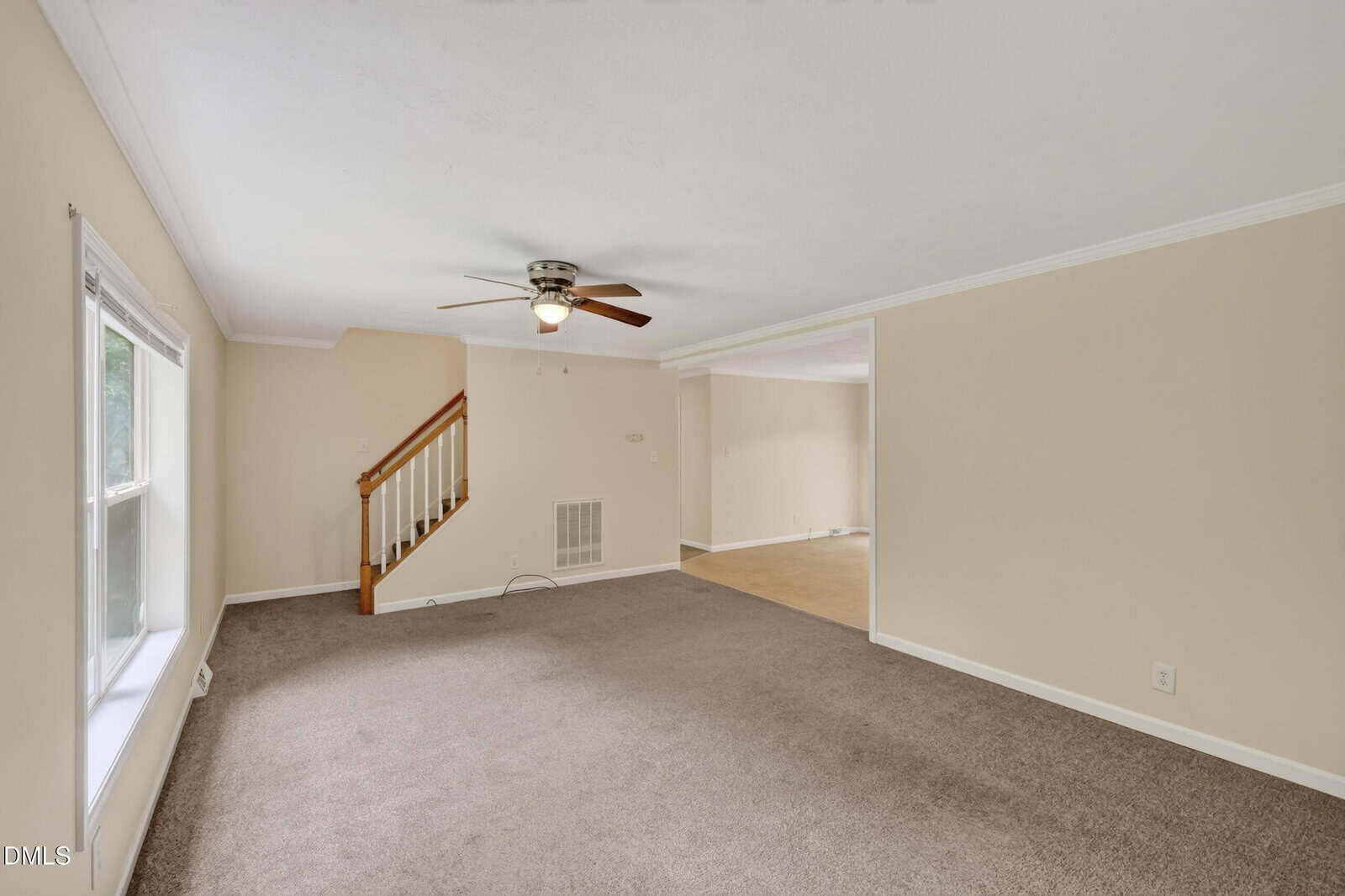 A spacious living room with beige walls, carpeted floor, a ceiling fan, and a window. A staircase with a wooden handrail leads upstairs, and an open doorway connects to another room with tile flooring.