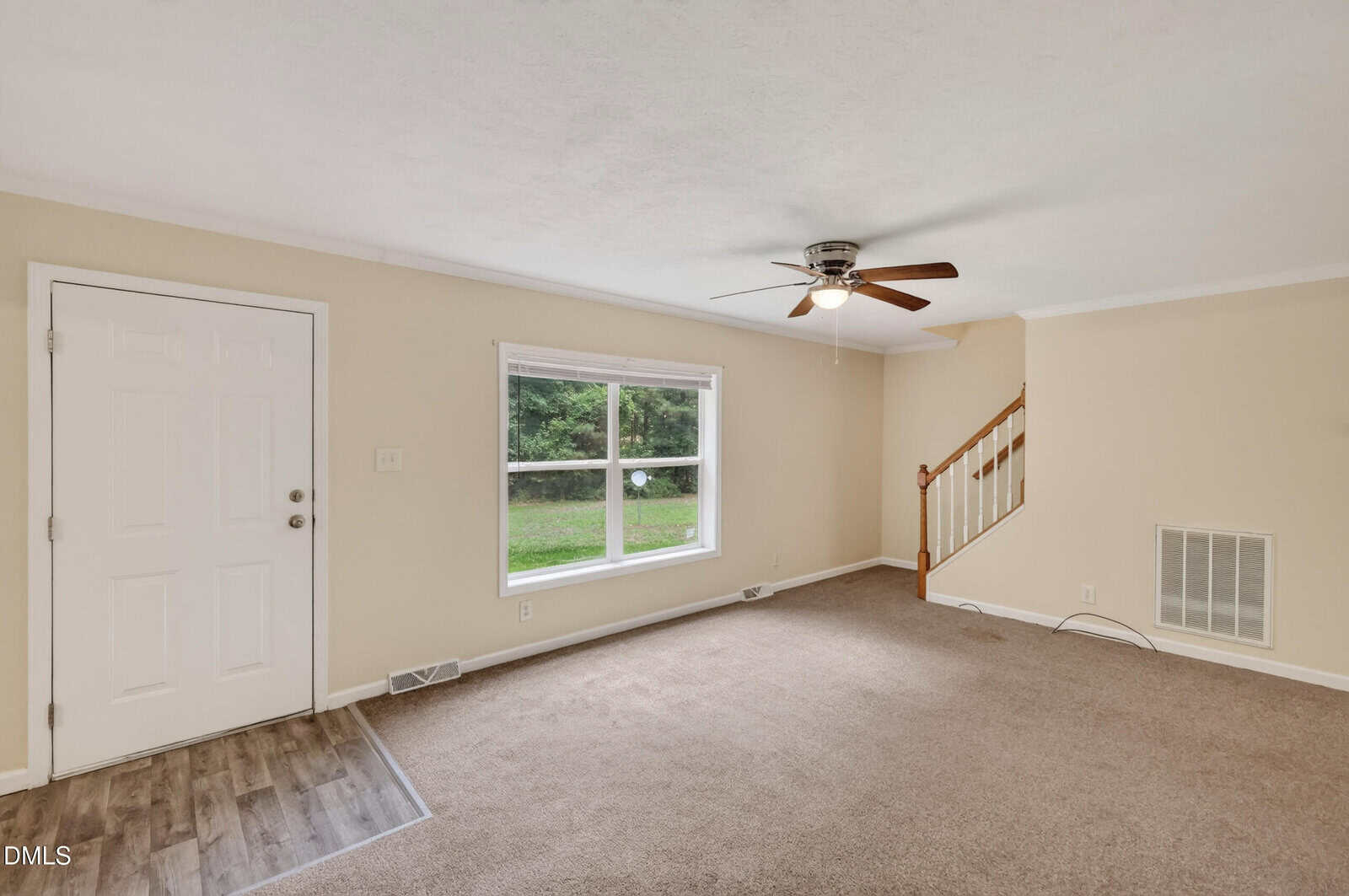 Bright living room with beige walls, carpeted floor, large window, ceiling fan, white front door, and stairs with a wooden handrail in the corner. Natural light fills the room.