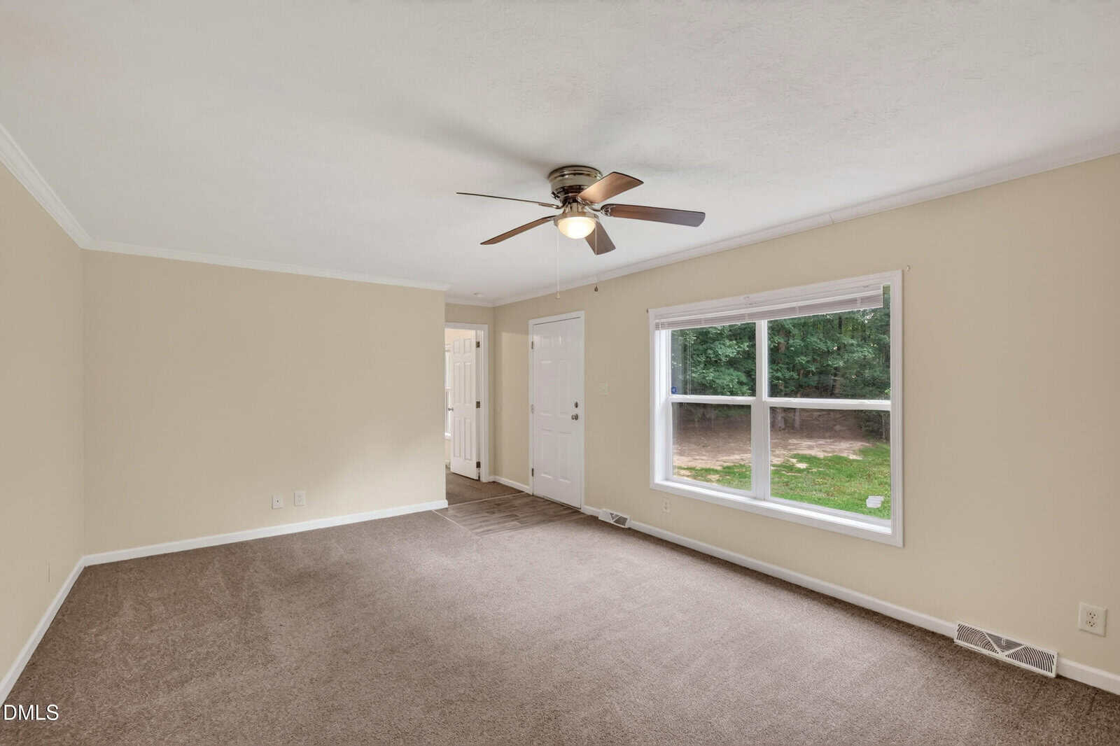 A spacious, empty room with beige walls, brown carpet, a ceiling fan with lights, a large window letting in natural light, and a white door leading outside.