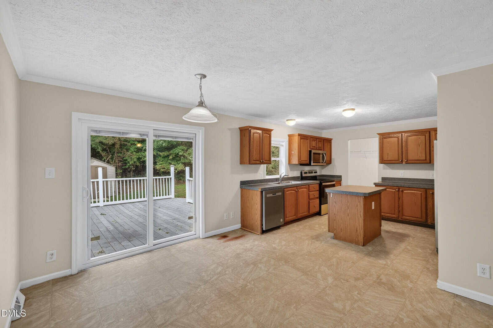 Kitchen with wood cabinets, stainless steel appliances, island, and adjoining dining area. Sliding glass doors open to a deck and green yard. Beige walls and light tile flooring throughout the space.