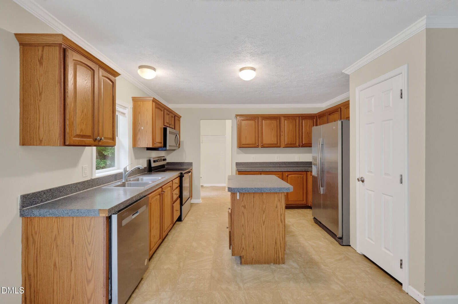 Spacious kitchen with wooden cabinets, stainless steel appliances, a central island, gray countertops, beige walls, and tiled flooring. Two ceiling lights provide illumination. Doorways lead to other rooms.