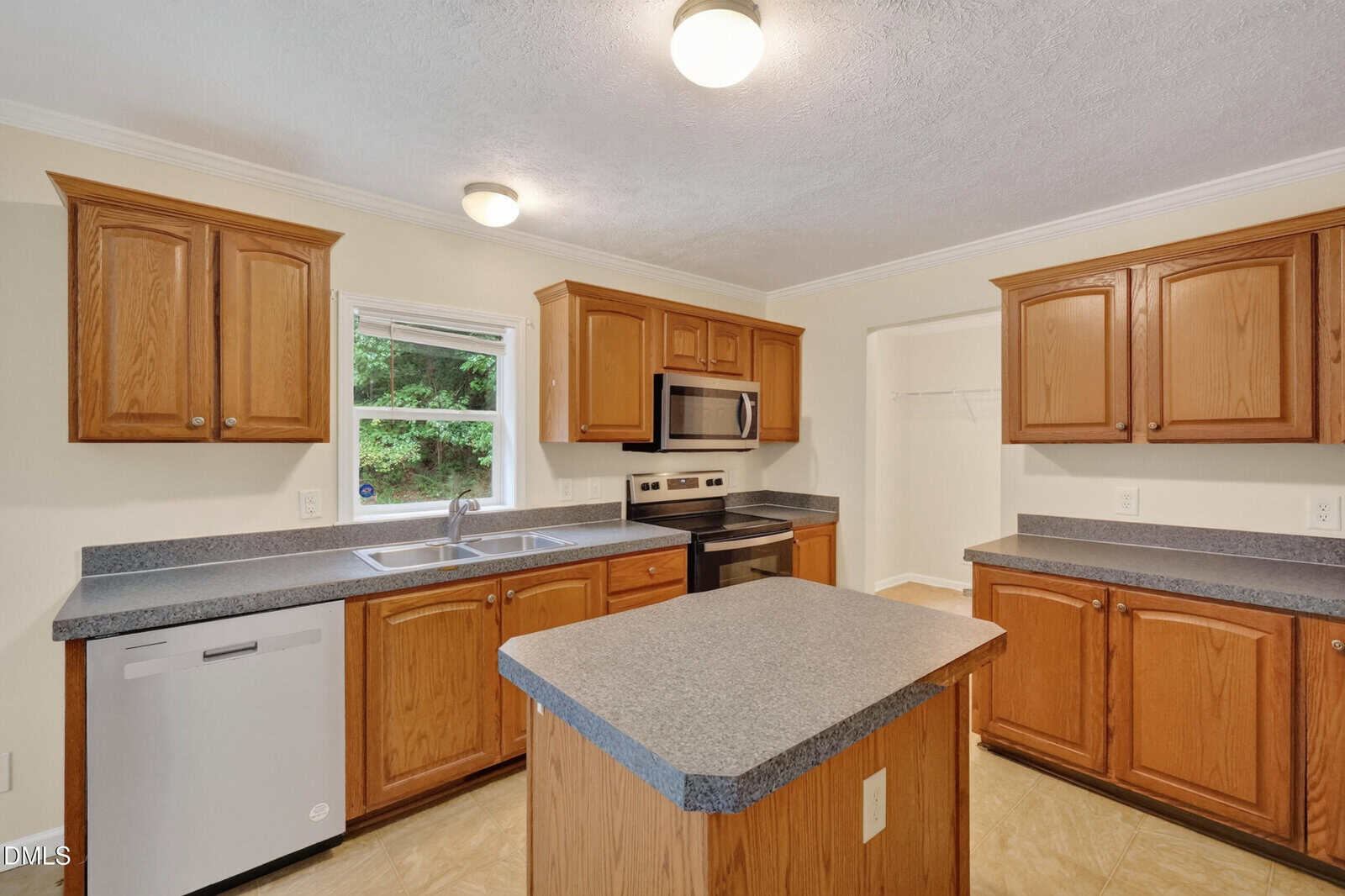 A kitchen with light brown wooden cabinets, gray countertops, a central island, stainless steel appliances, a double sink under a window, and light-colored tile flooring. Ceiling lights illuminate the space.