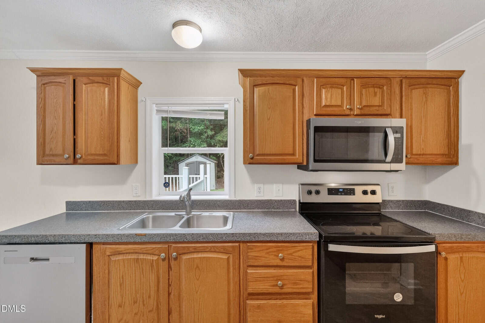 A kitchen with light wood cabinets, gray countertops, a double sink, stainless steel dishwasher, oven, and microwave. A window above the sink shows green trees and part of a white outdoor structure.