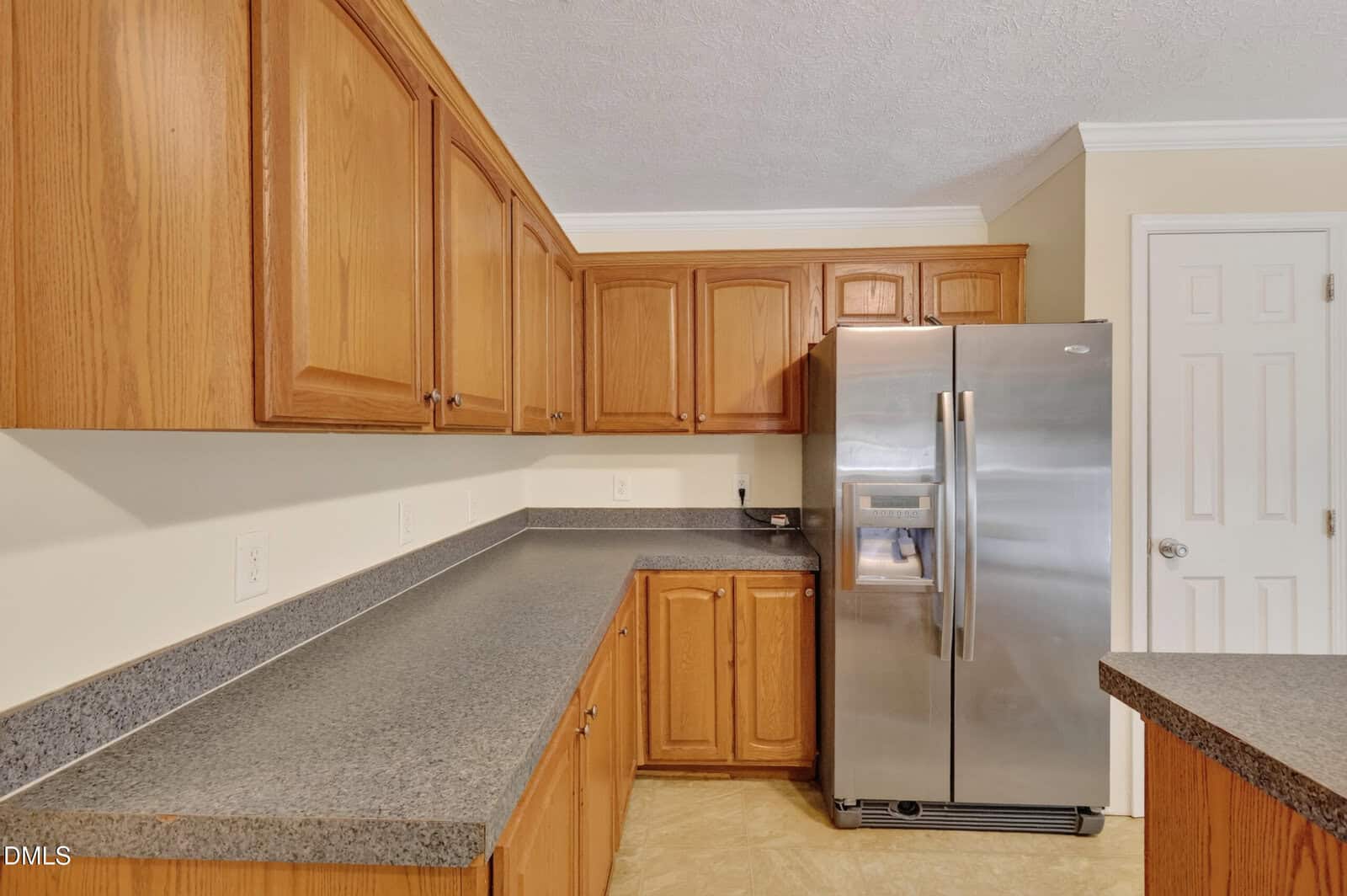 Kitchen with wooden cabinets, gray speckled countertops, a stainless steel side-by-side refrigerator, light beige walls, and a closed white door in the background.
