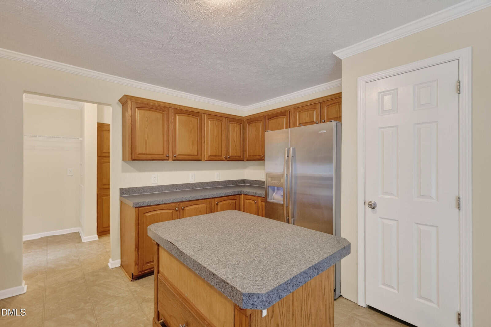 Kitchen with wood cabinets, gray countertops, central island, stainless steel refrigerator, and beige walls. A white door and open doorway are visible. Light tan tile floor throughout the space.