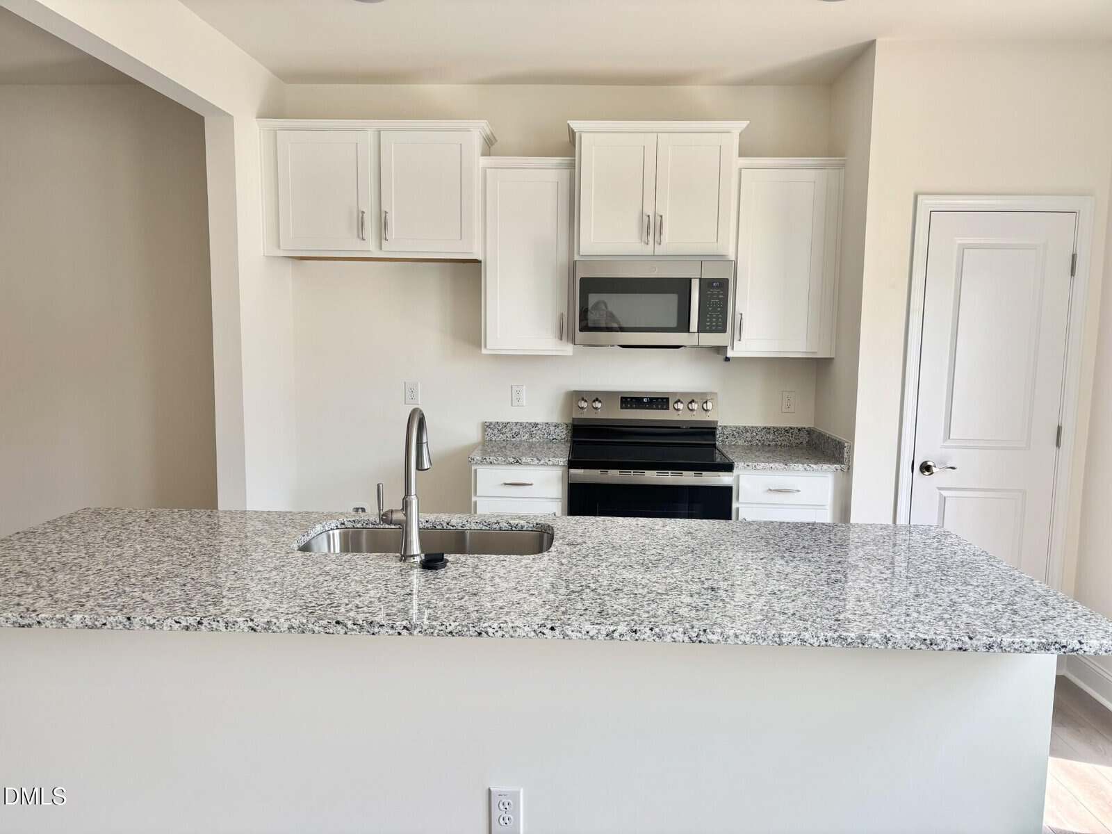 Modern kitchen with white cabinets, granite countertops, stainless steel stove and microwave, sink on island, and a closed white door to the right. The space is bright with neutral walls.