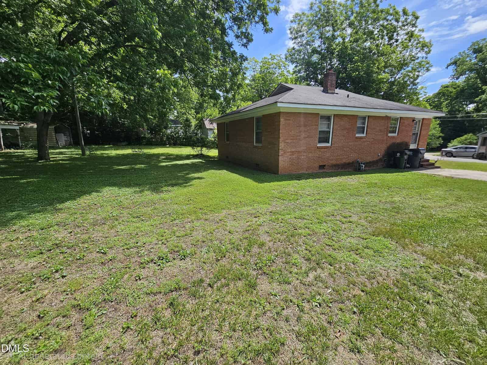 A brick house with a gray roof sits on a spacious grassy yard, surrounded by large green trees under a partly cloudy blue sky. Trash bins are next to the house and a driveway is visible on the right.