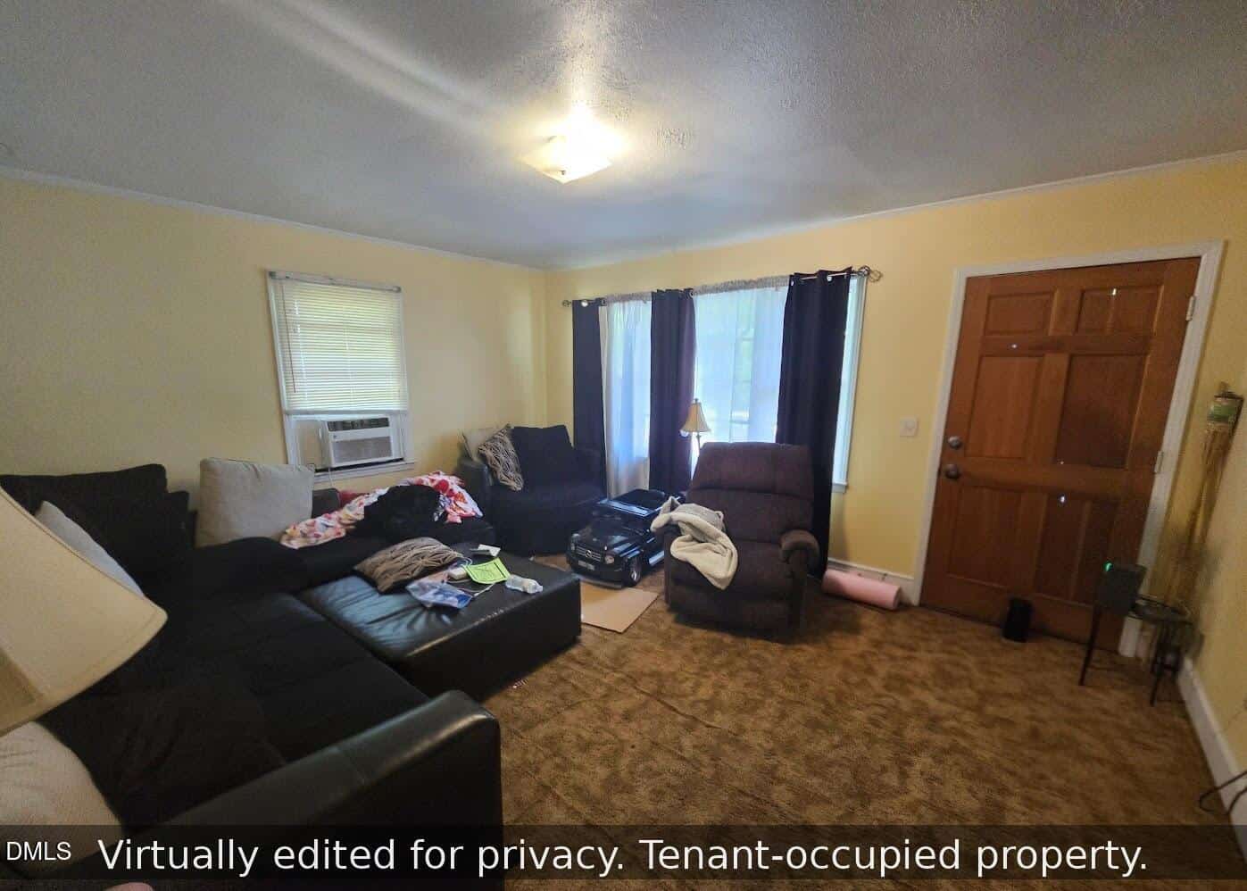 Living room with brown carpet, sectional sofa, recliner, scattered clothes, suitcases, and a lamp. Two windows with curtains, an air conditioner, and a wooden front door are visible. Text overlay notes tenant-occupied property.