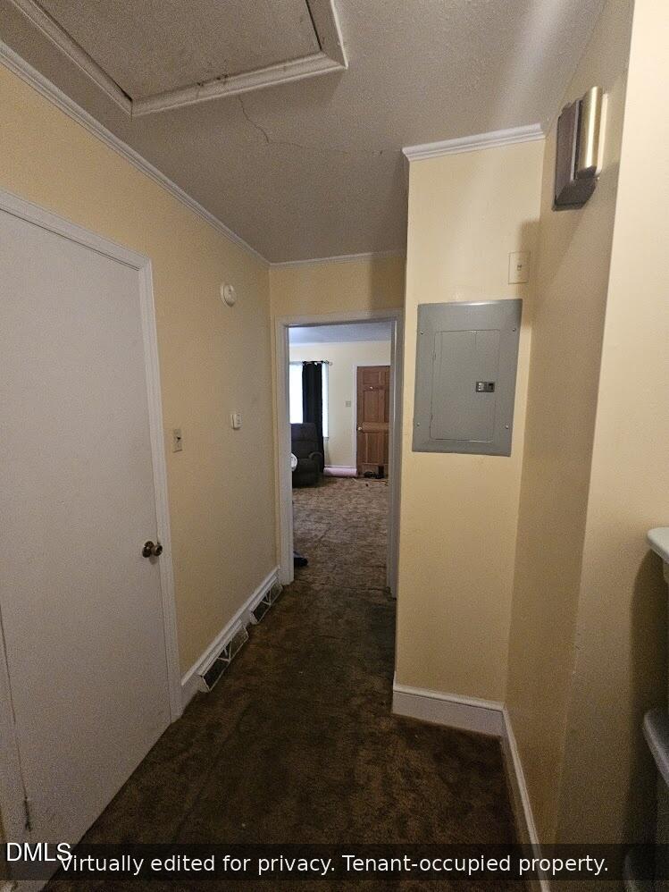 A hallway with beige walls and brown carpet, featuring a closed white door on the left, an electrical panel on the right wall, and a room visible at the end with natural light and a wooden door.