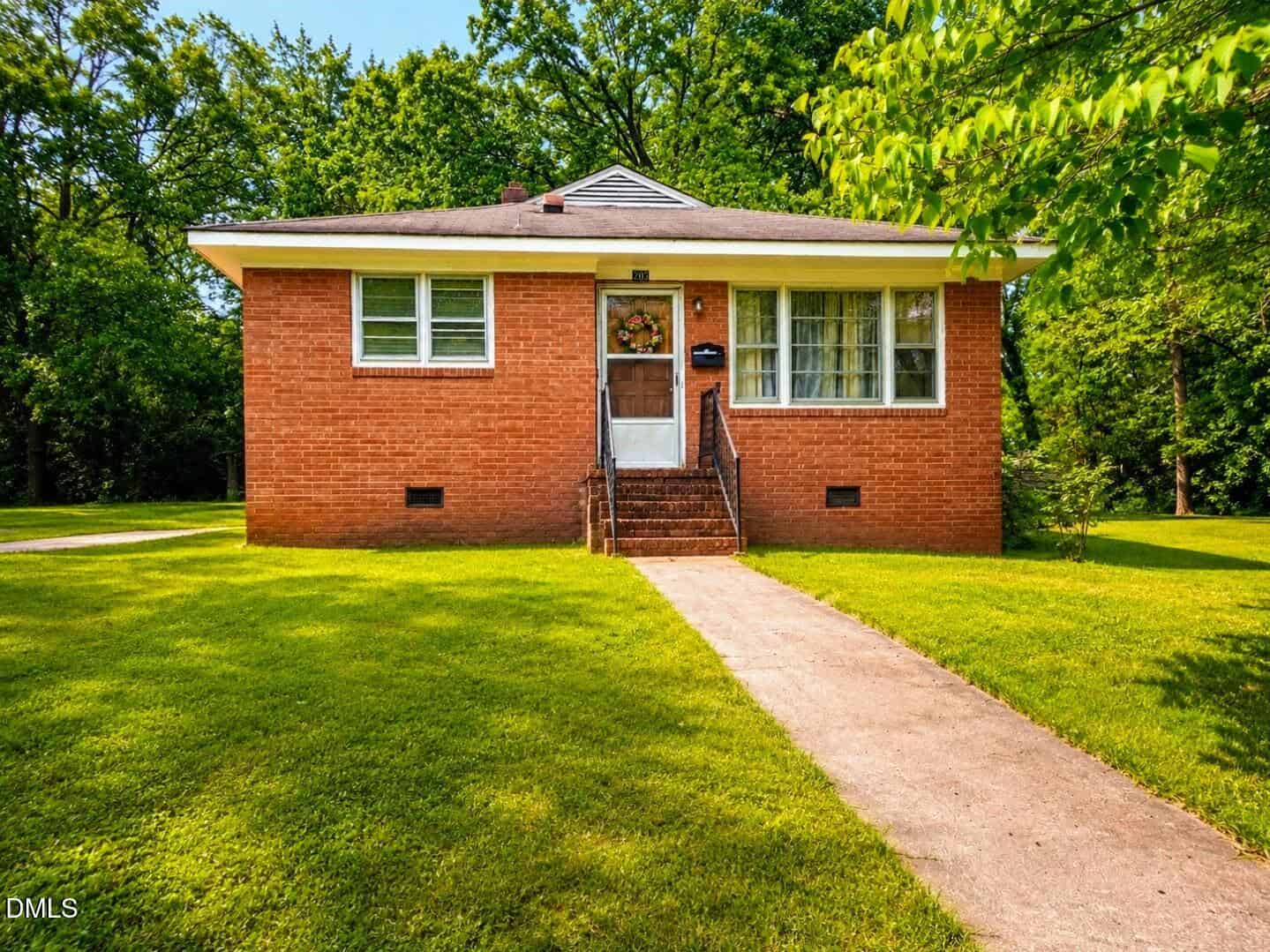 A small, single-story brick house at 209 Lee Street, Oxford, features a grassy lawn, concrete walkway and steps to the front door. Large windows flank the entrance; trees and greenery surround the home on a bright, sunny day.