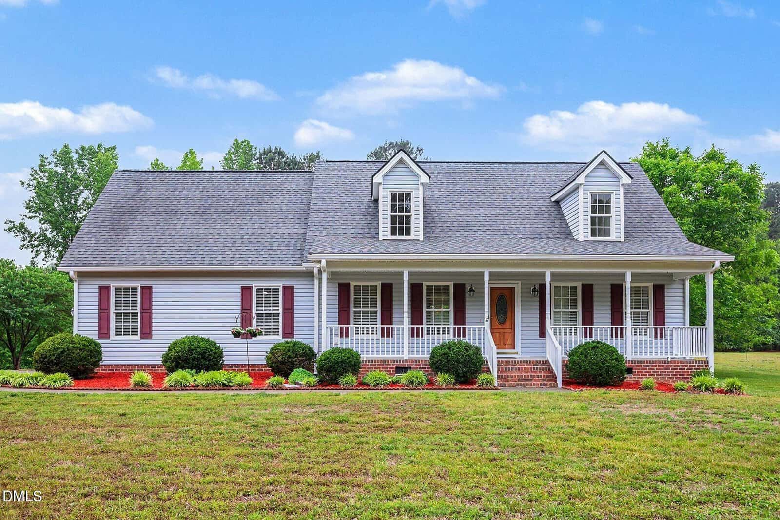 A charming one-story house at 100 Linnett Court, Youngsville, featuring gray siding, red shutters, and a spacious front porch with white railings. The yard boasts trimmed bushes, a lush lawn, mature trees, and a blue sky backdrop.
