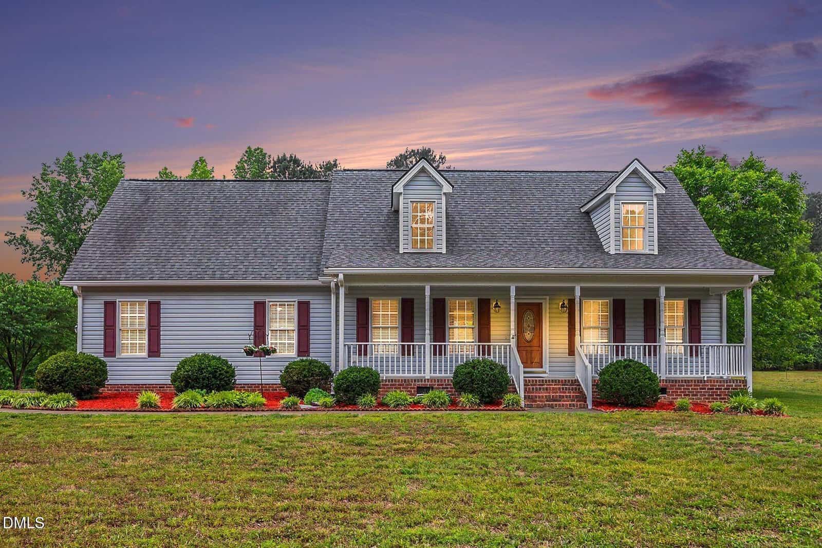 A charming gray house with a large front porch, white trim, and dormer windows, surrounded by manicured bushes and a green lawn at sunset.
