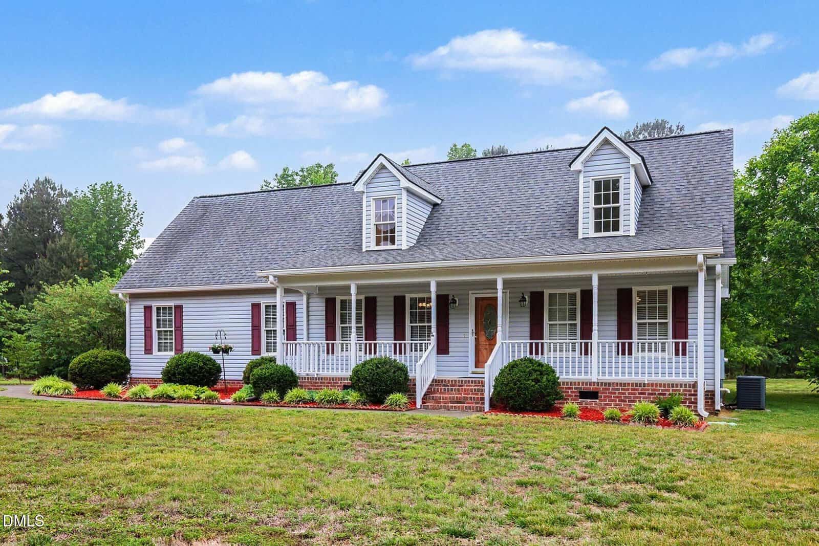 A white, single-story house with a large front porch, brick steps, dormer windows, and burgundy shutters, surrounded by neatly trimmed bushes, red mulch, and a grassy lawn under a blue sky with scattered clouds.
