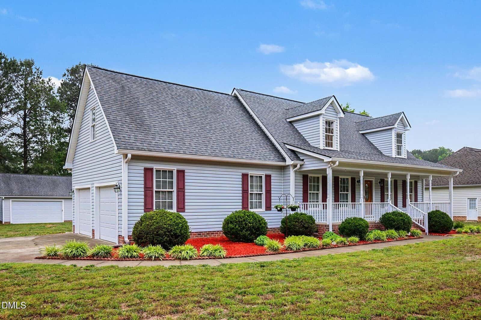 A single-story house with gray siding, dark red shutters, a large front porch, dormer windows, manicured bushes, and landscaped flower beds in the front yard under a clear blue sky.