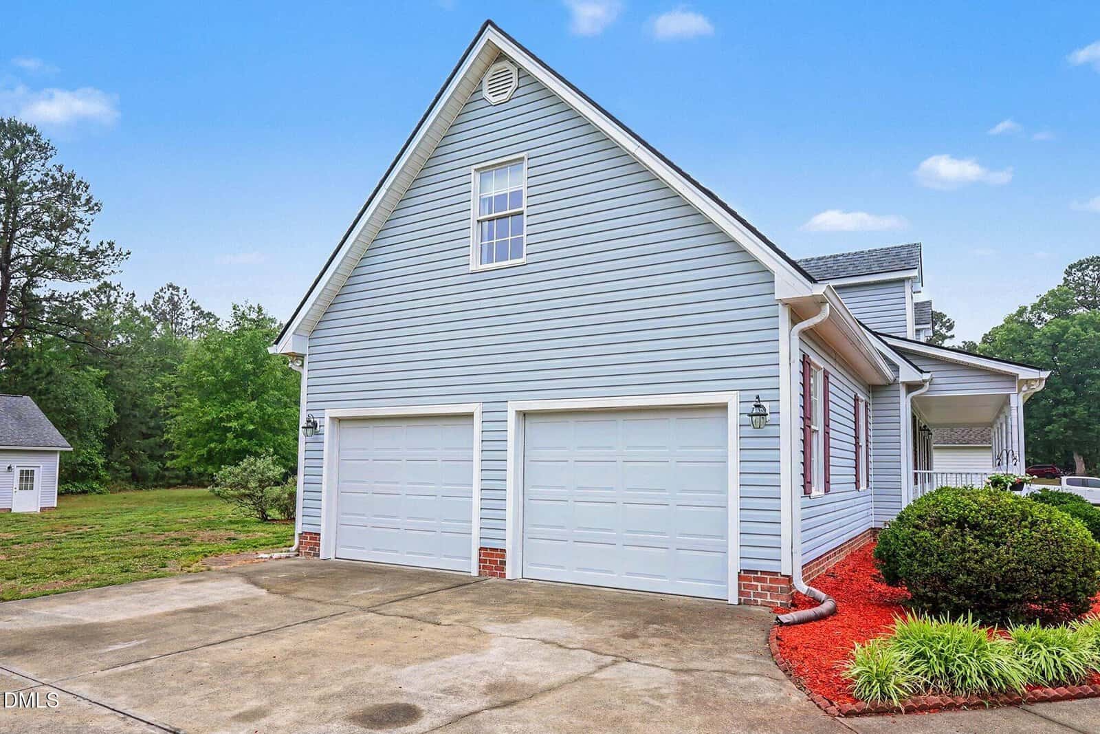 A light blue house with white trim features a double garage and a large driveway. Red mulch and bushes line the right side, and a grassy yard with trees is in the background under a blue sky with clouds.
