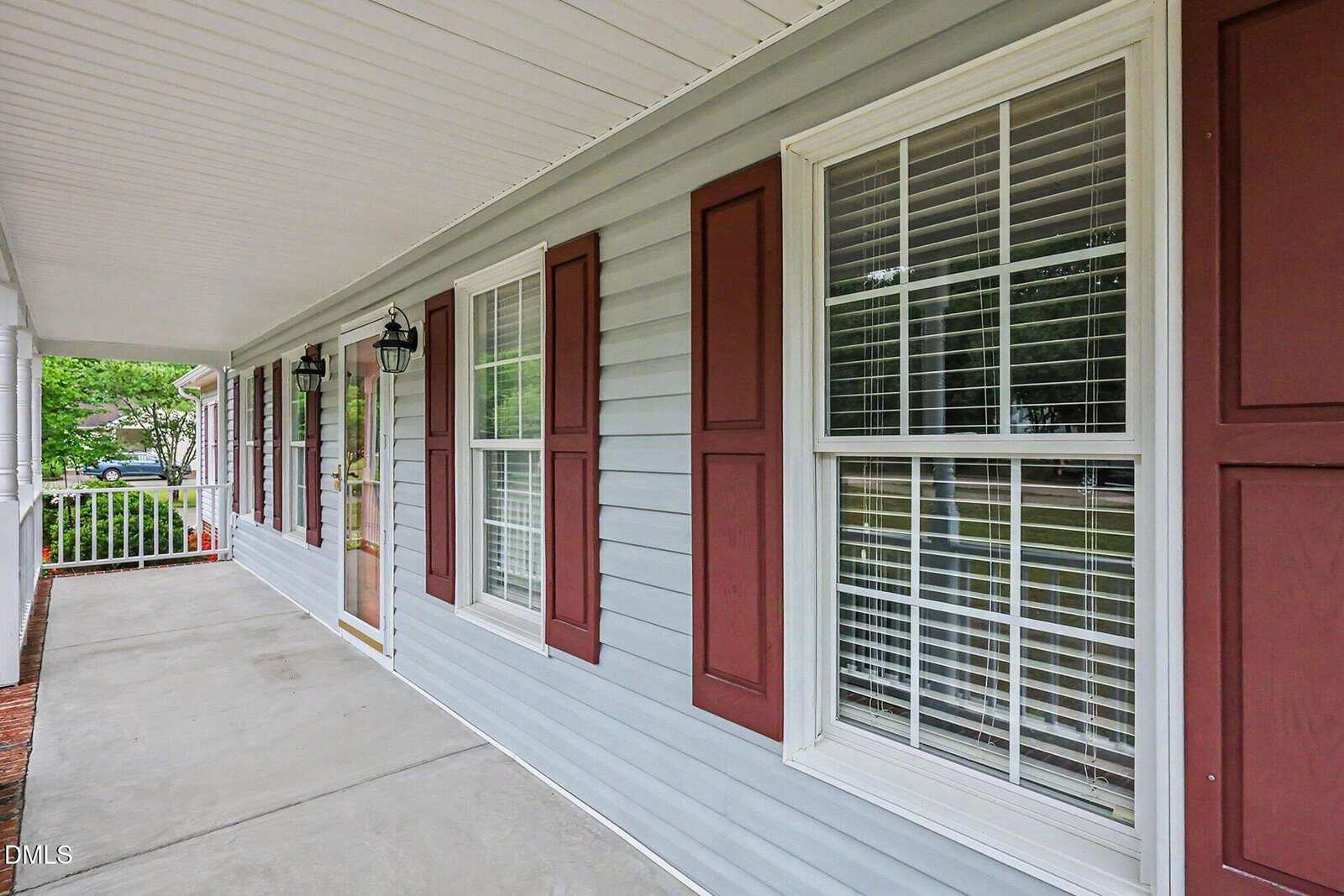 A gray house exterior with white trim, maroon shutters, large windows, a covered porch, and a concrete walkway. Two black lantern-style lights are mounted by the front door. Trees and greenery are visible in the background.