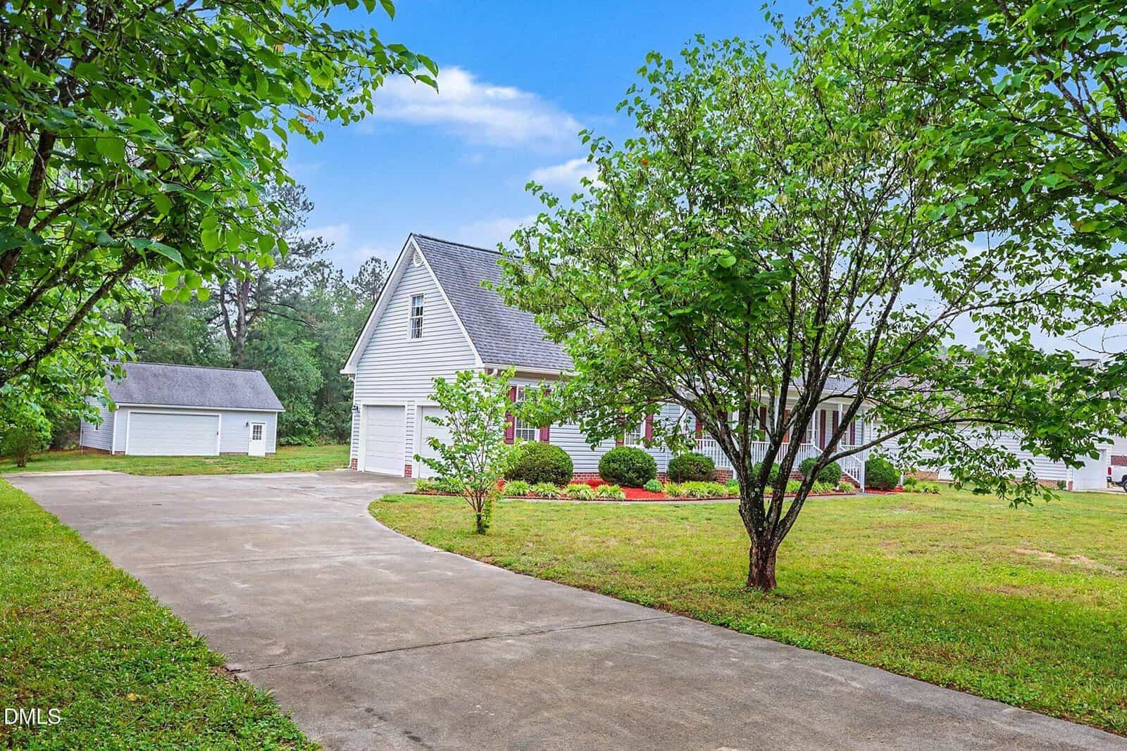 A white house with a front porch and attached garage sits beside a curved driveway. Green trees and a detached white garage are in the yard under a blue sky.