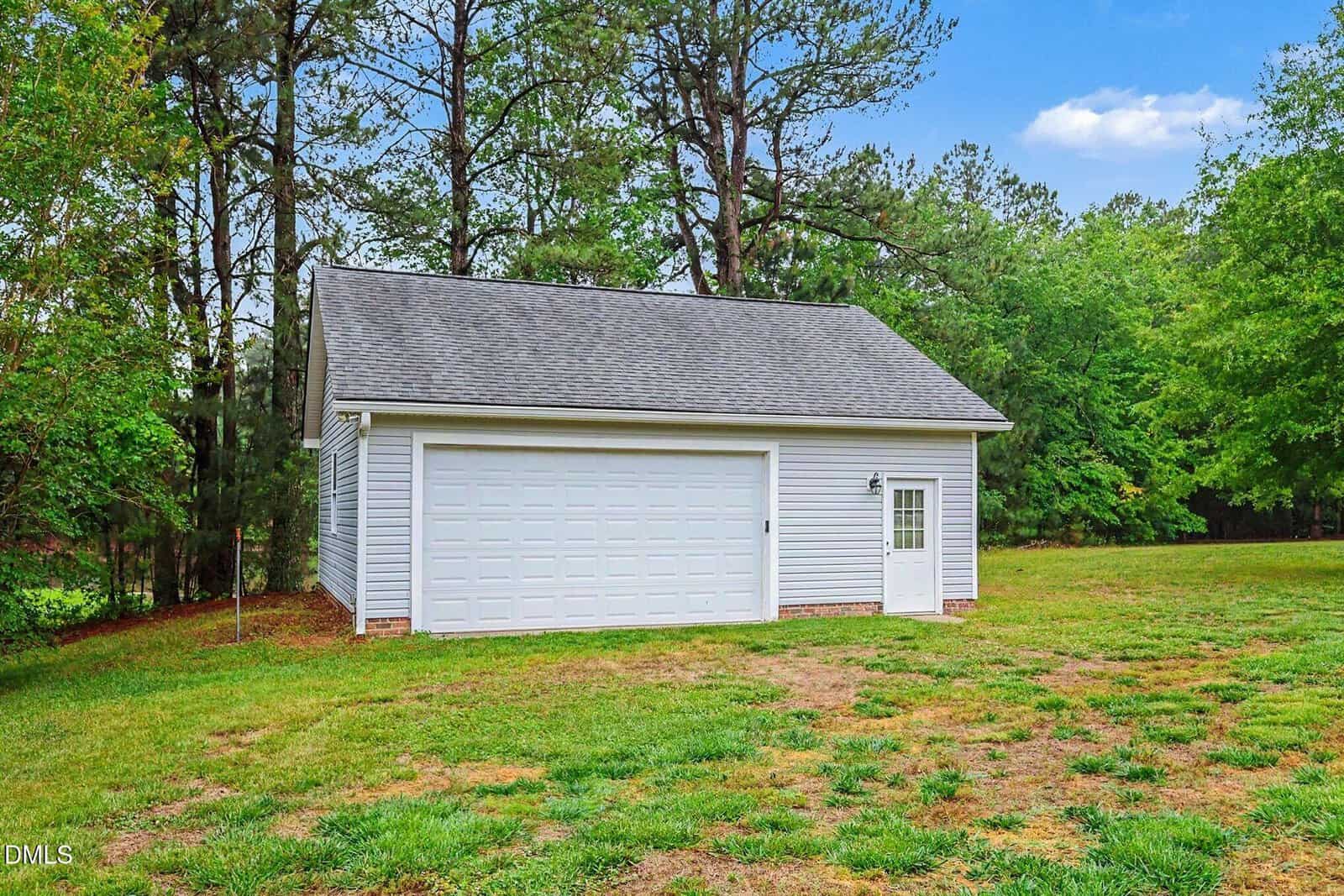 A detached gray garage with a white door and side entry door, surrounded by grass and trees, under a blue sky with some clouds.